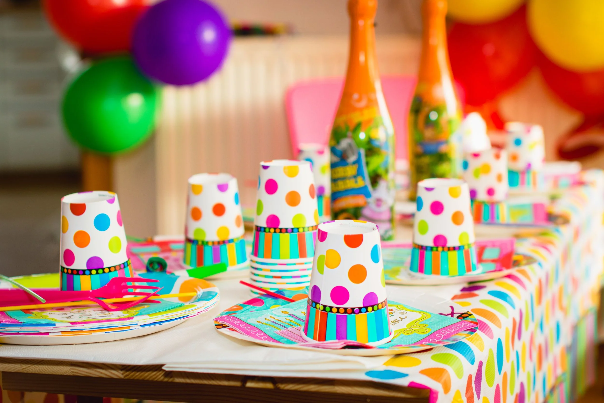 Colorful birthday party table decorated with polka dot paper cups, vibrant paper napkins, and party plates, with balloons in the background.