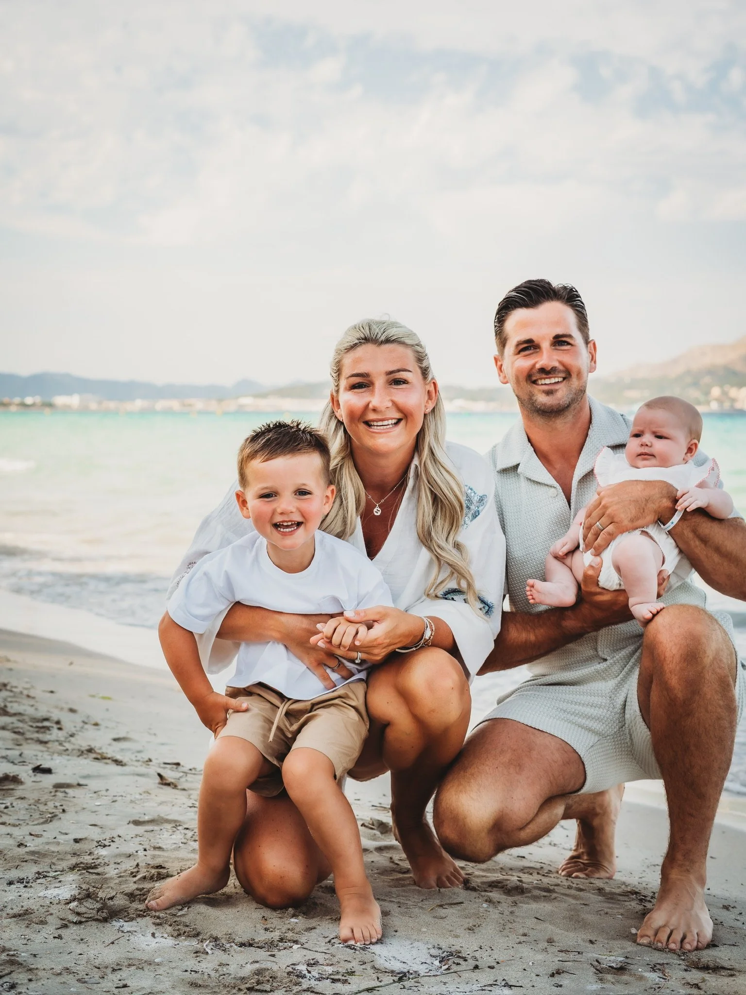 A family of four at the beach, smiling and posing for the photo. The father is holding a baby girl, the mother has two children, a young boy and girl, with the beach and water in the background.