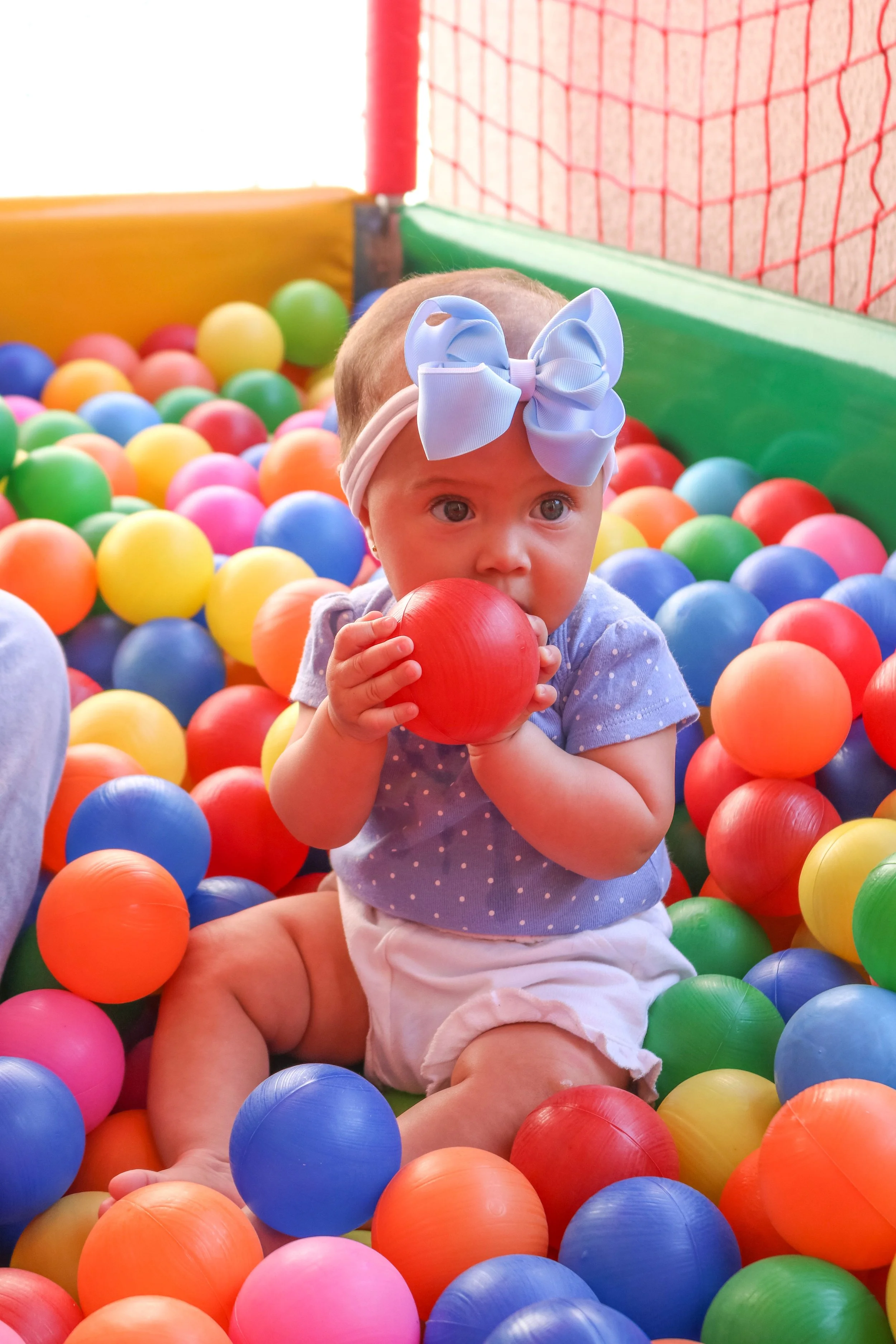 Young girl with big blue bow headband playing in a colorful ball pit, biting a red ball, surrounded by orange, yellow, pink, blue, green balls.