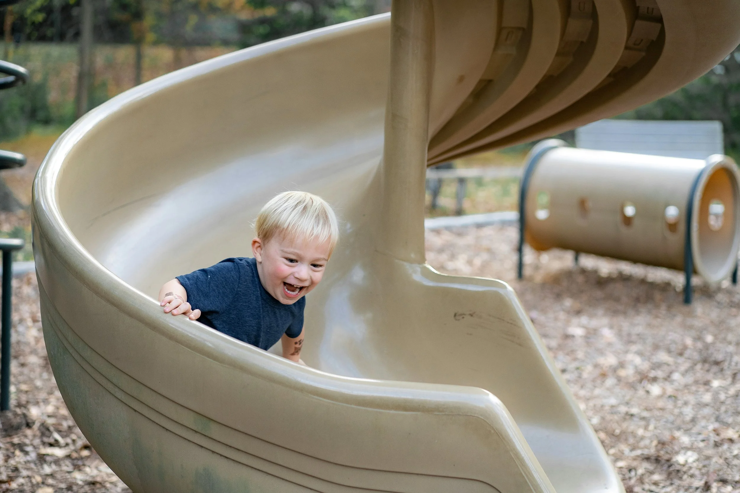 A young boy with blonde hair and a blue shirt smiling as he slides down a beige playground slide at a park during daytime, surrounded by fallen leaves and trees.