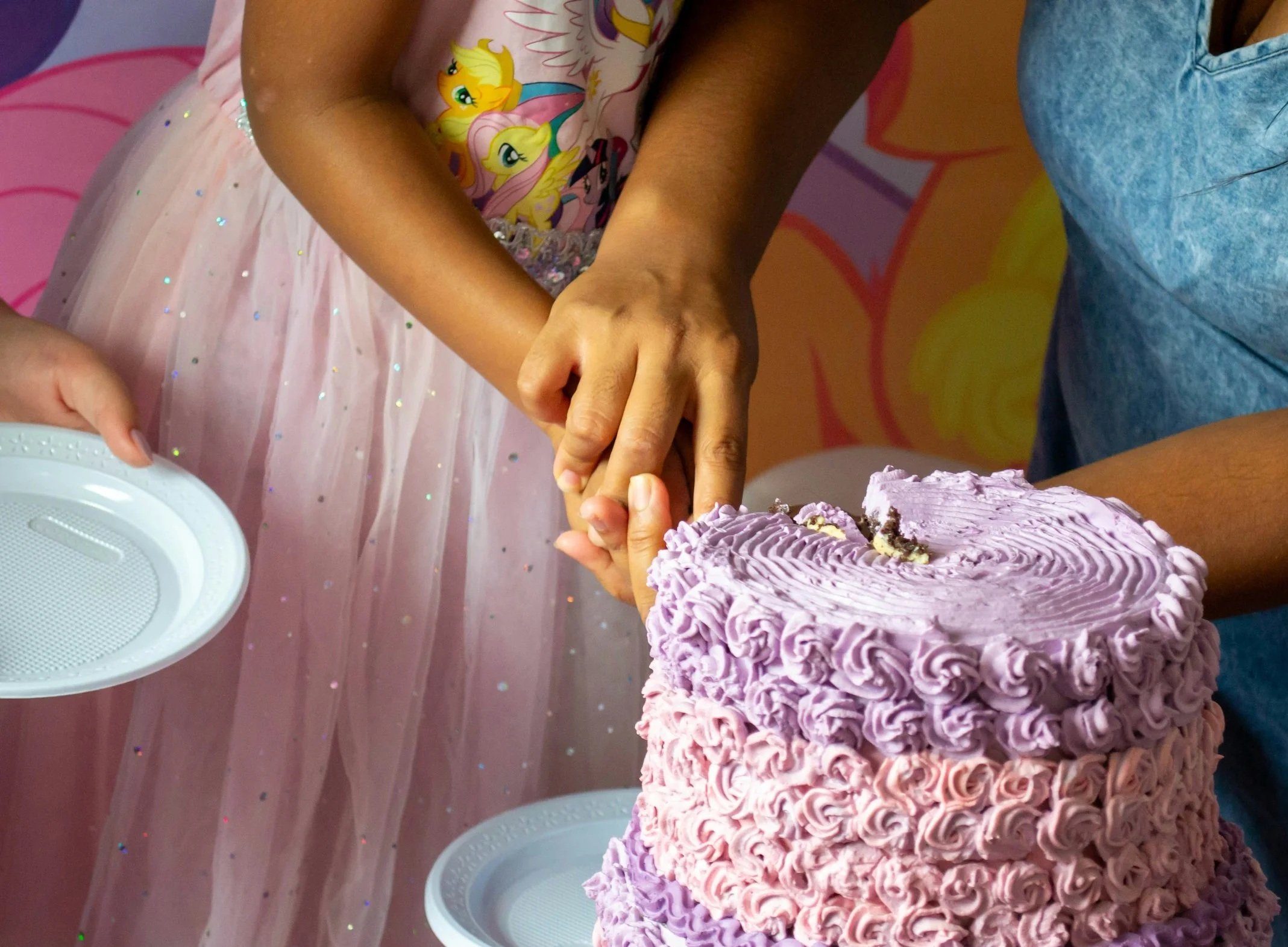 Two children are holding each other's hands while standing near a birthday cake with purple frosting. The children are wearing colorful clothing, one with a pink