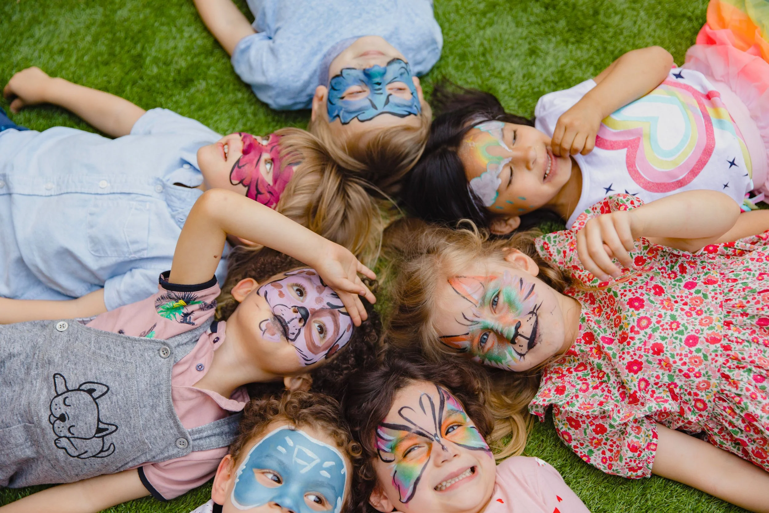 Children lying on grass with face paint, smiling and looking up, wearing colorful clothes.