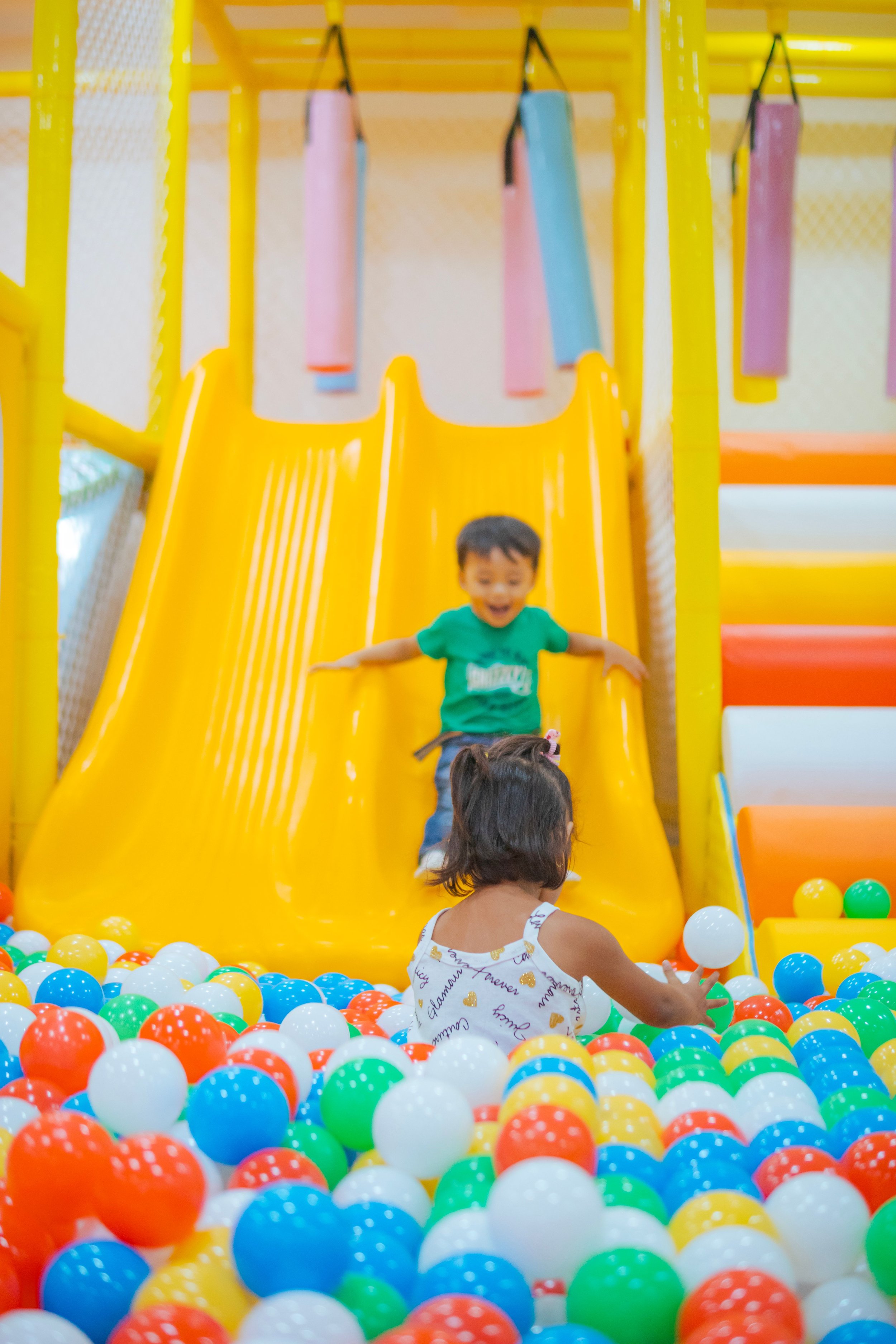 A child sliding down a yellow slide into a ball pit with colorful plastic balls, while another child reaches into the ball pit and a woman watches.