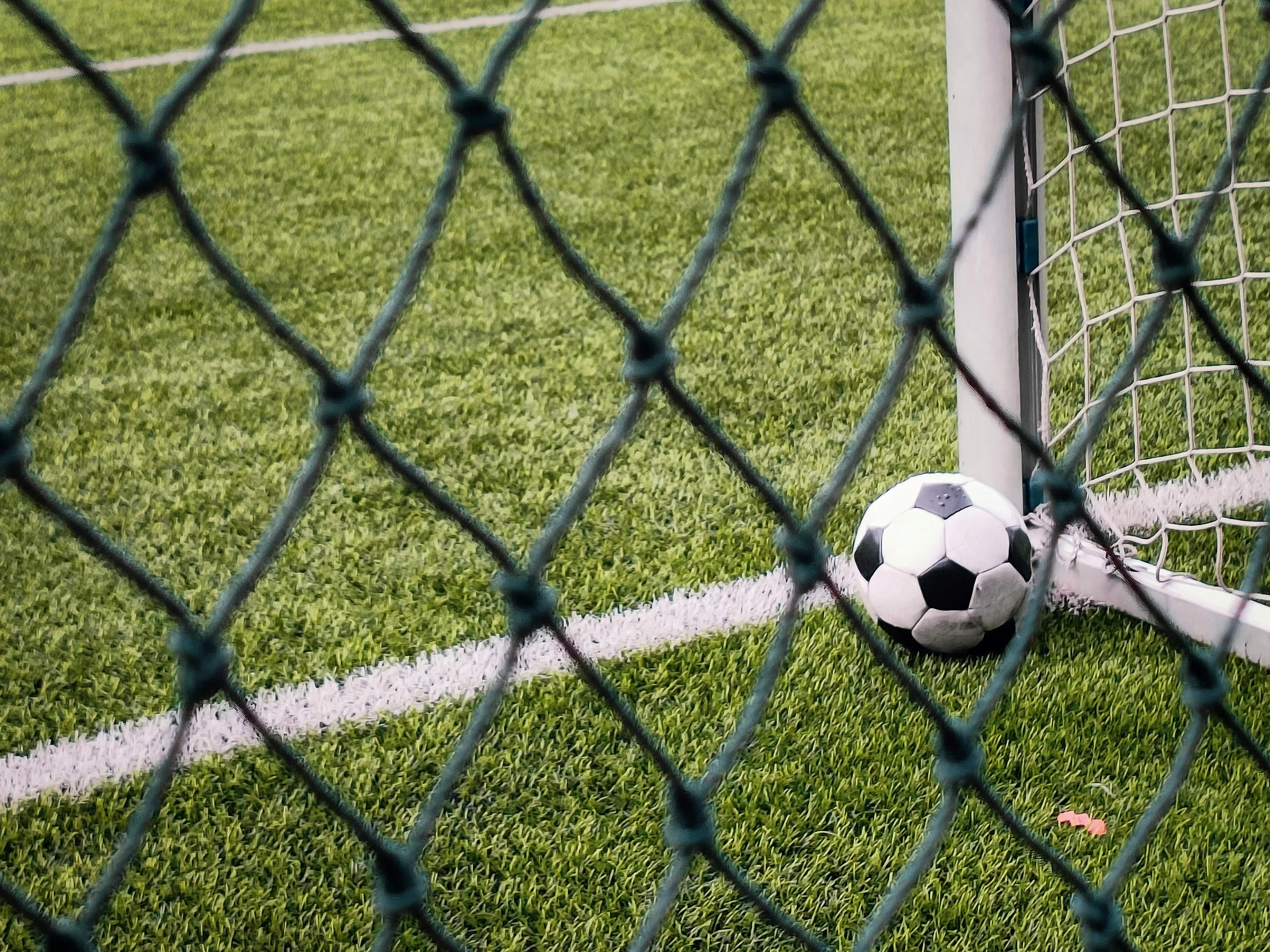 A soccer ball resting near the goalpost inside a soccer field, viewed through a chain-link fence.