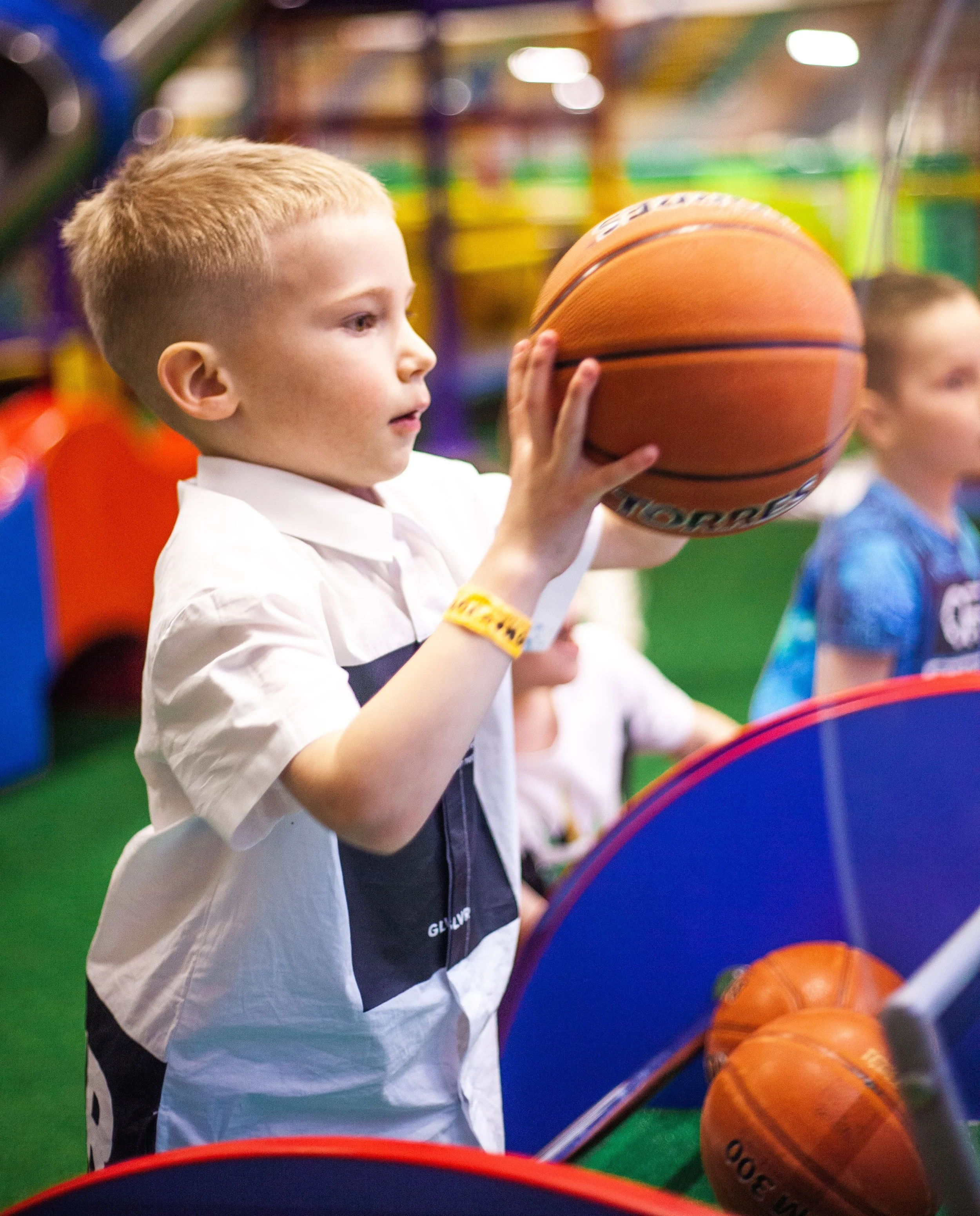 Young boy with short blond hair holding a basketball inside an arcade or entertainment center.