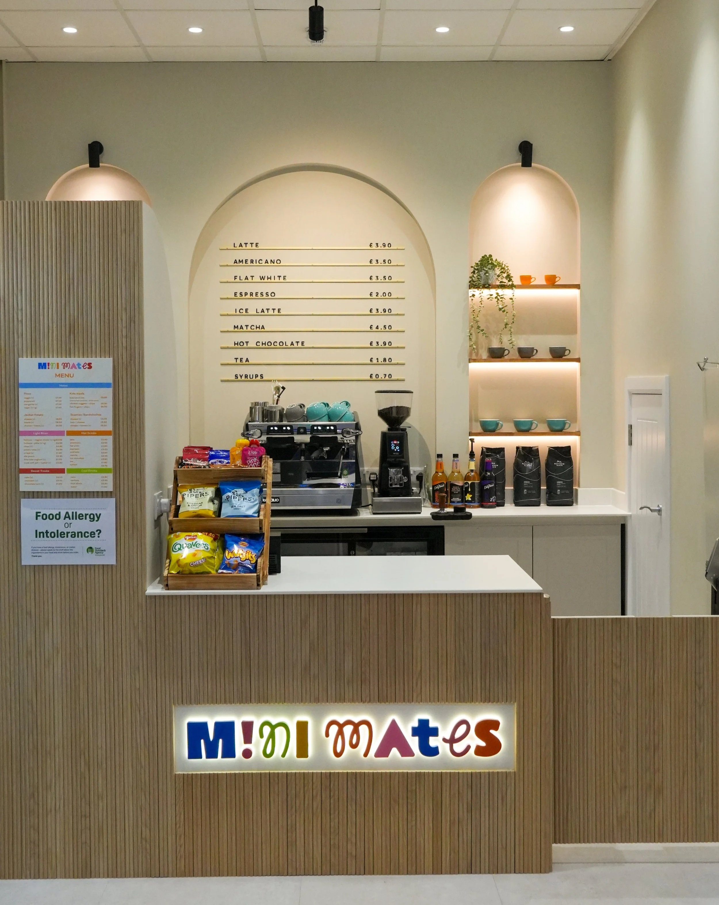 Interior of a modern coffee shop with large arched windows, hanging pendant lights, a white wall menu board displaying coffee options, and a counter with cups and equipment. Customers are seated near the windows.