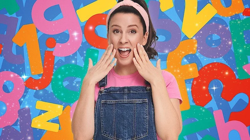 Young woman with dark hair wearing a pink headband and pink shirt under denim overalls, surprised expression with hands on cheeks, colorful background with large multicolored numbers and sparkles.
