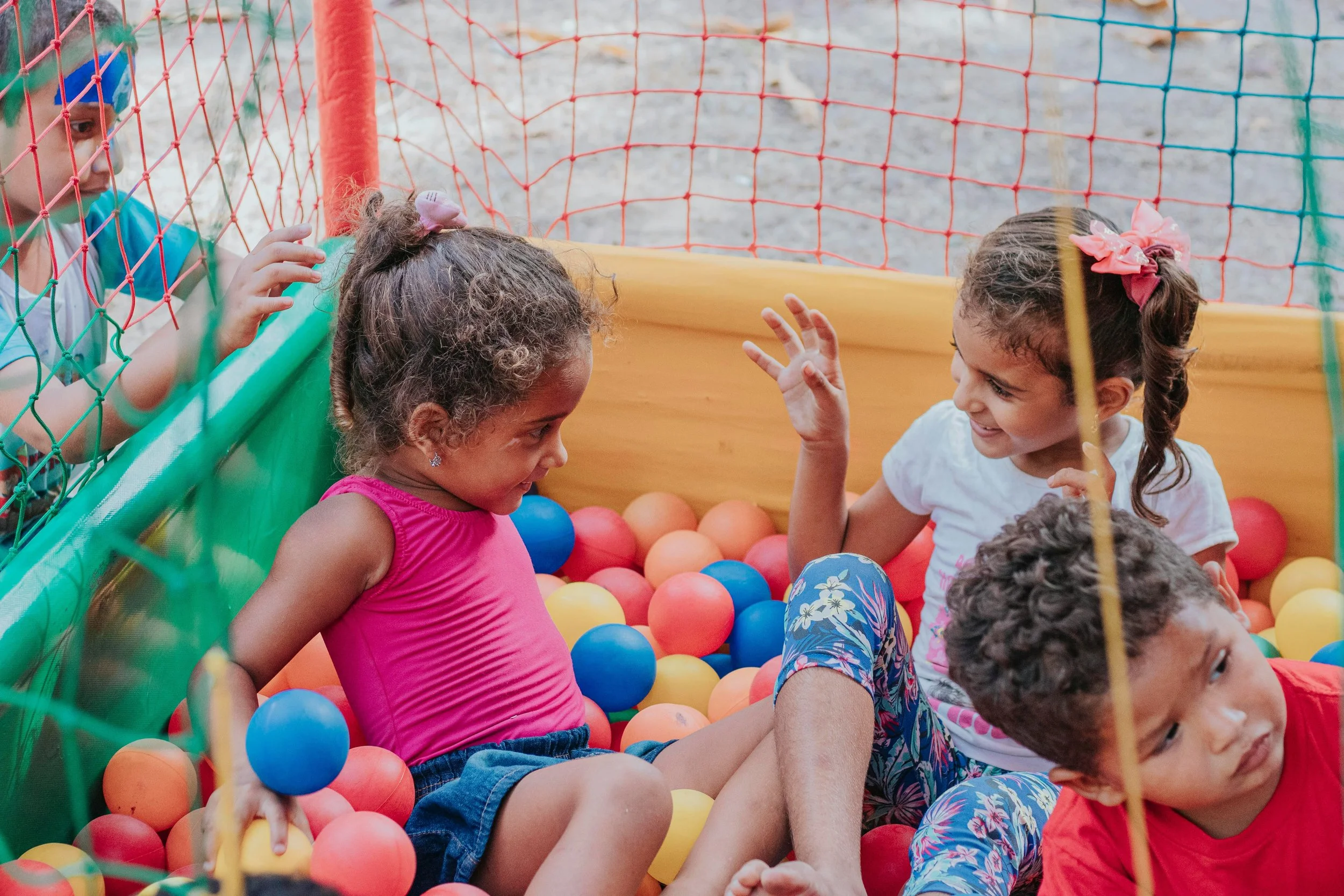 Children playing and sitting in a ball pit at a playground, behind a colorful safety net.