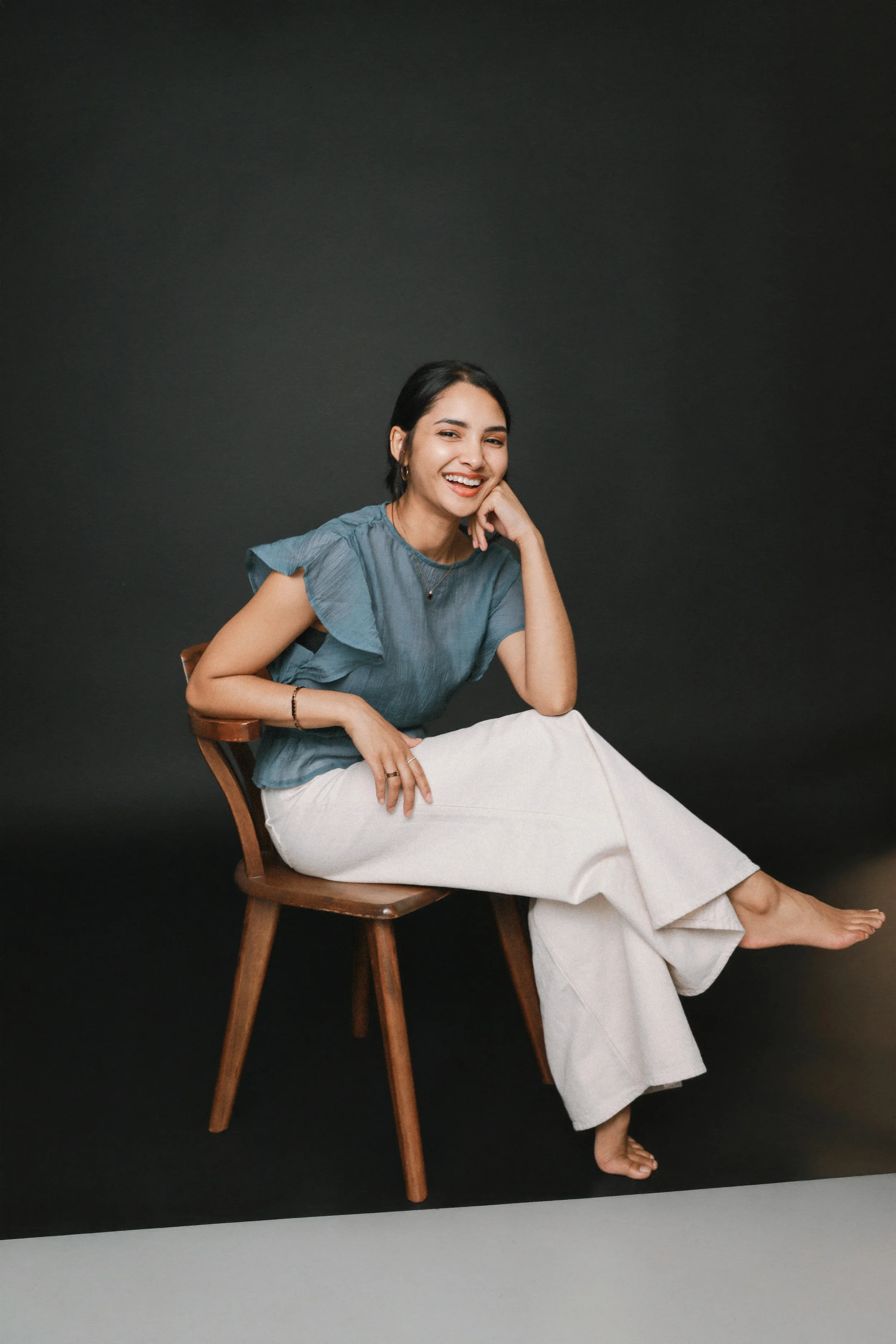 A woman sitting on a wooden chair with a dark background. She is smiling, wearing a blue top with ruffled sleeves, white wide-legged pants, and has short black hair and earrings.