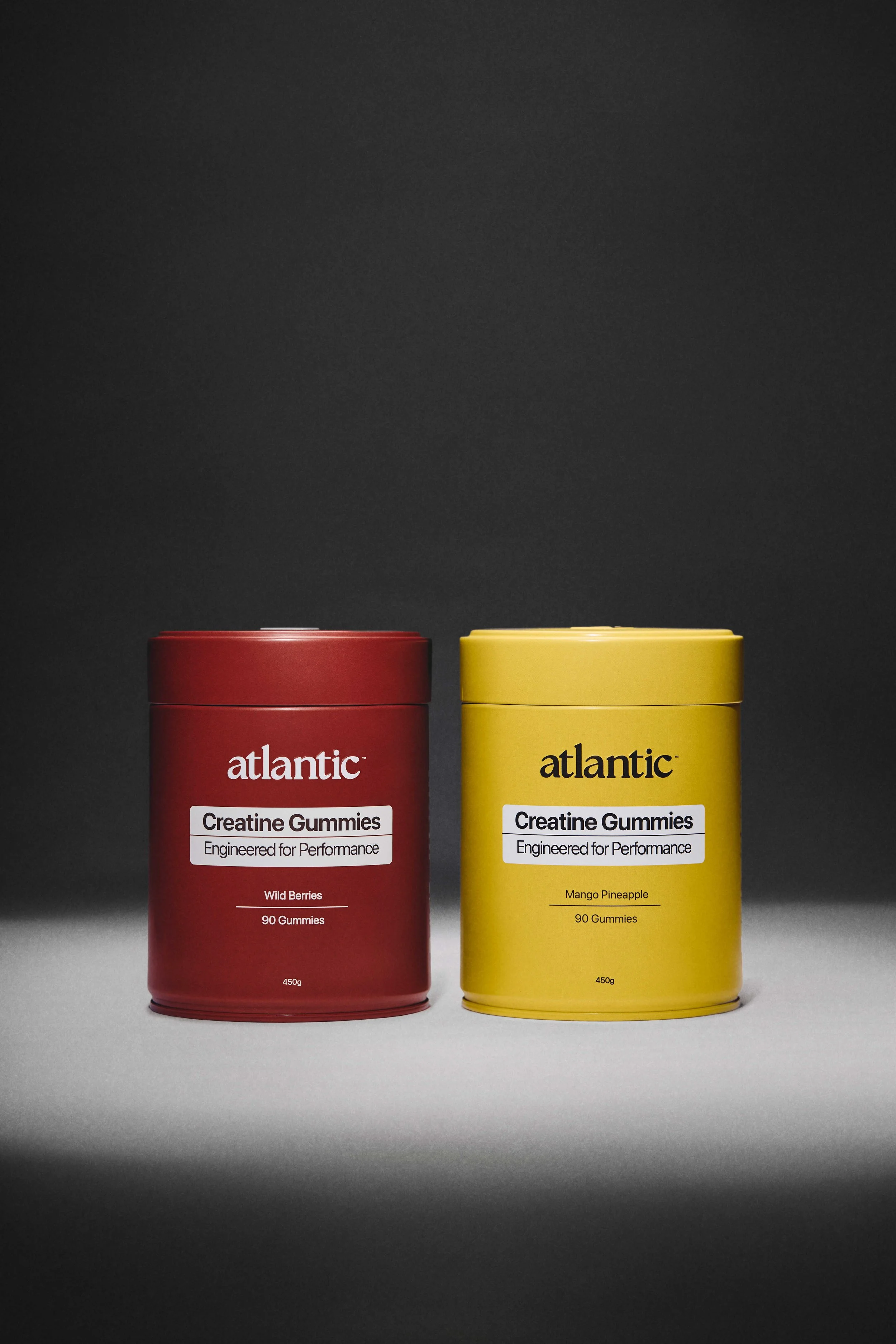 Two cylindrical containers of Atlantic Creatine Gummies, one red with wild berries flavor and one yellow with mango pineapple flavor, placed against a dark background.