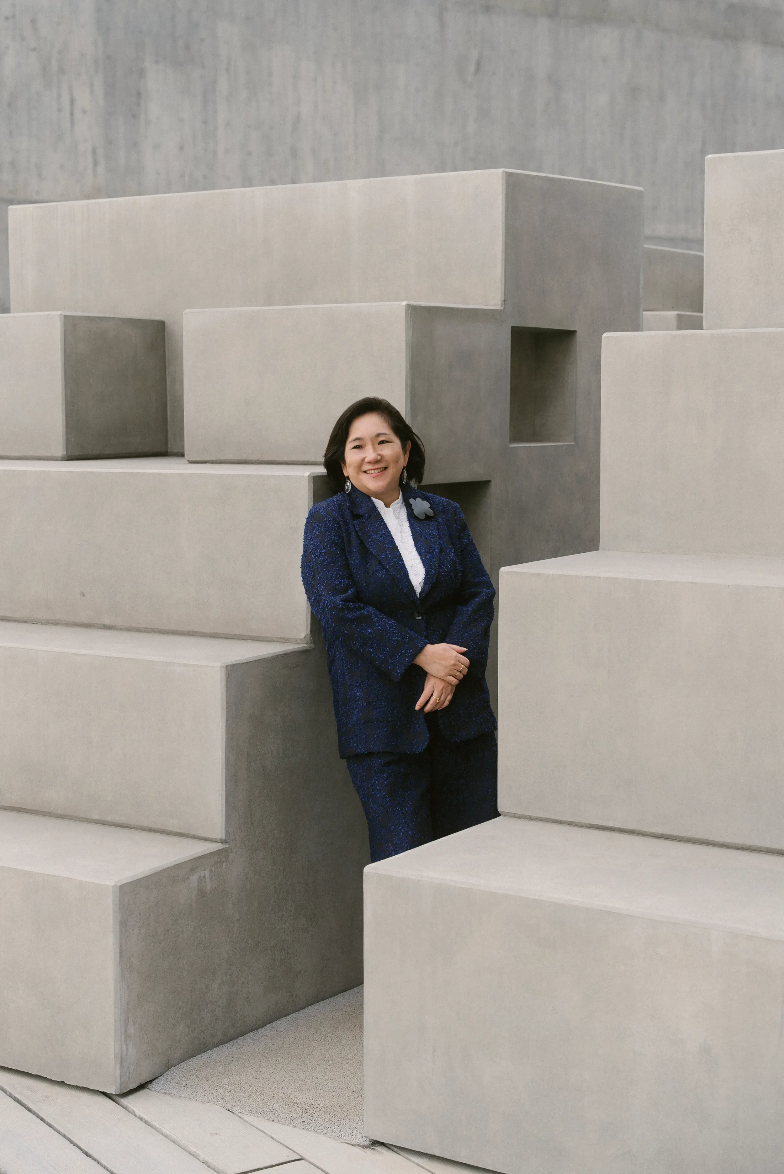 A woman with dark hair, wearing a blue patterned blazer, white shirt, and earrings, standing and smiling among large, modern, gray concrete blocks.