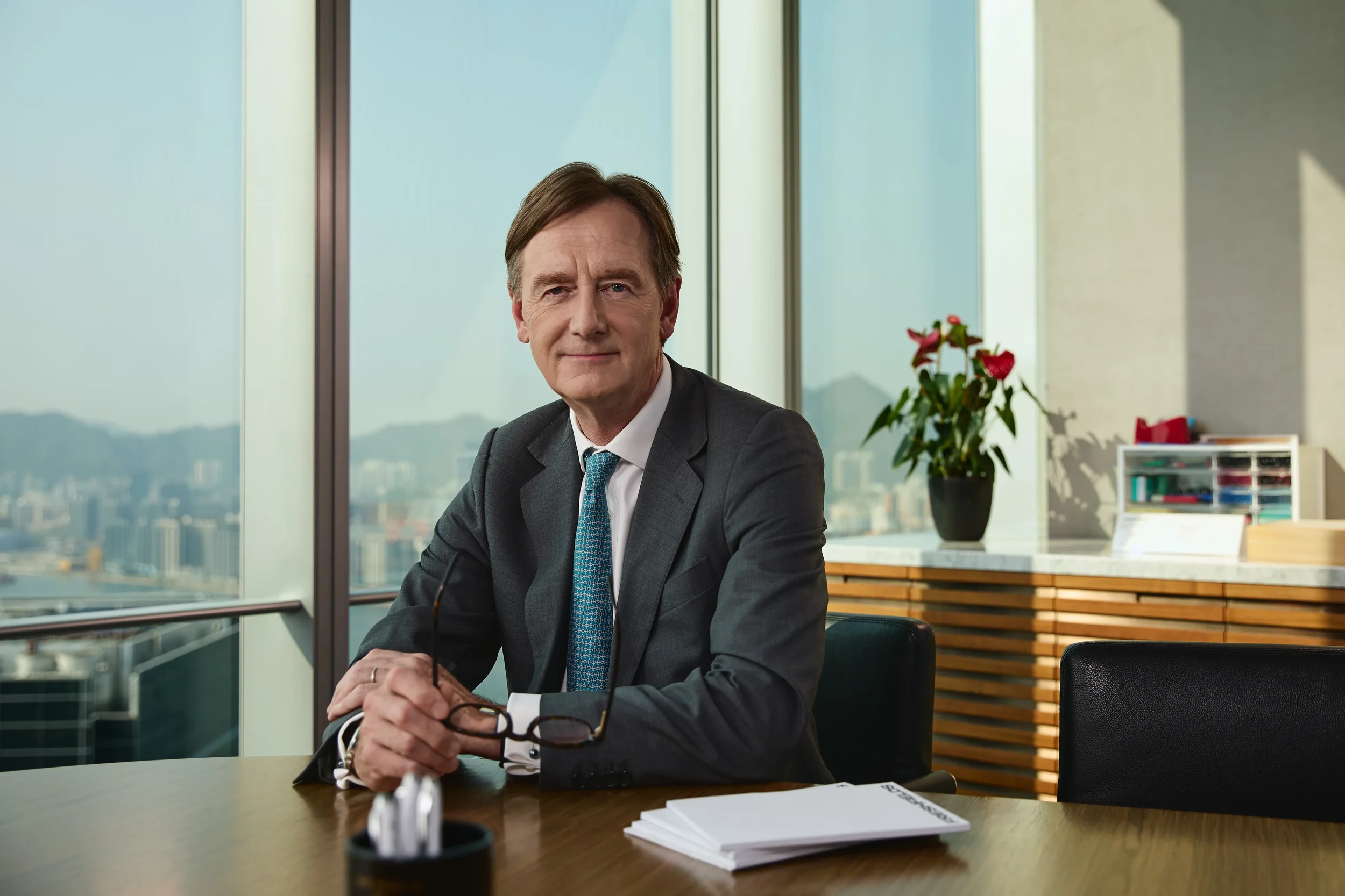 A middle-aged man in a gray suit and tie sitting at a conference table in an office with large windows showing a city skyline and mountains in the background. He is holding glasses in his hands, with a stack of papers and a pen in front of him, and a