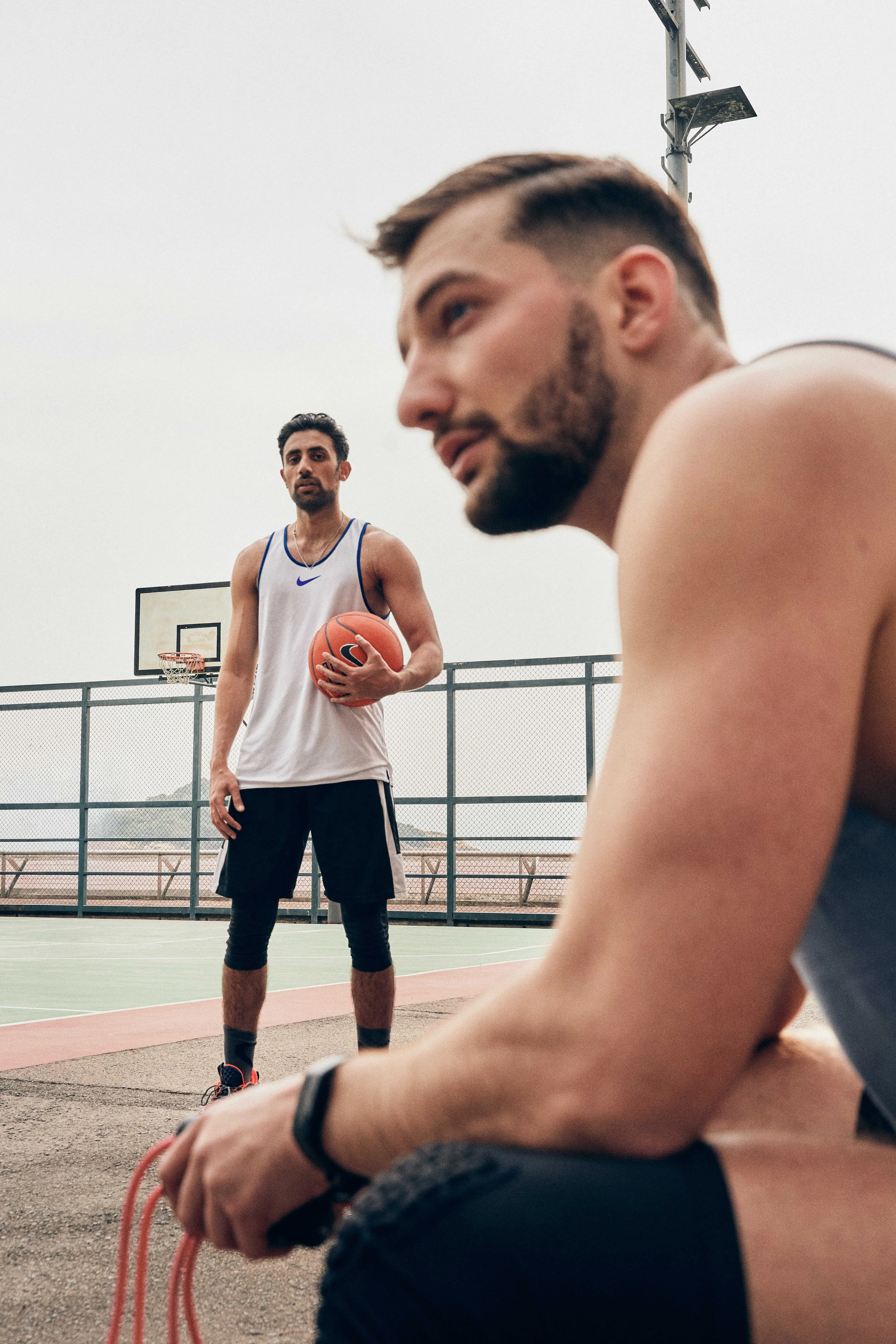 Two men on an outdoor basketball court on an overcast day: one holding a basketball in the background, the other in close-up foreground holding a jump rope.