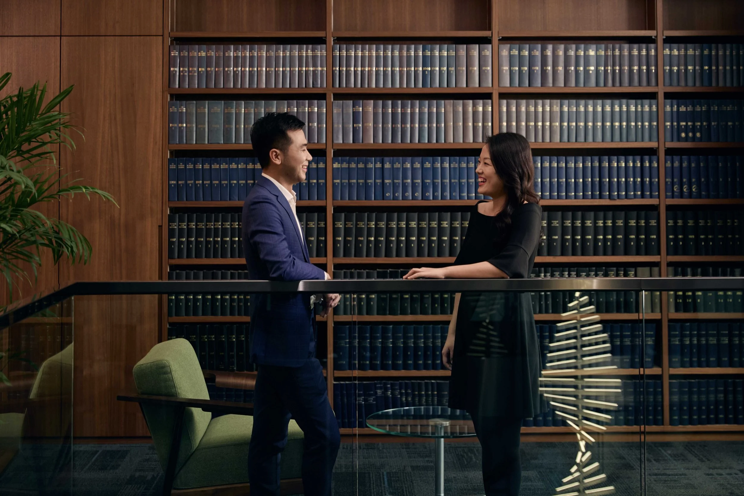 A man and woman standing and smiling at each other in an office or library with wooden bookshelves filled with blue books behind them.