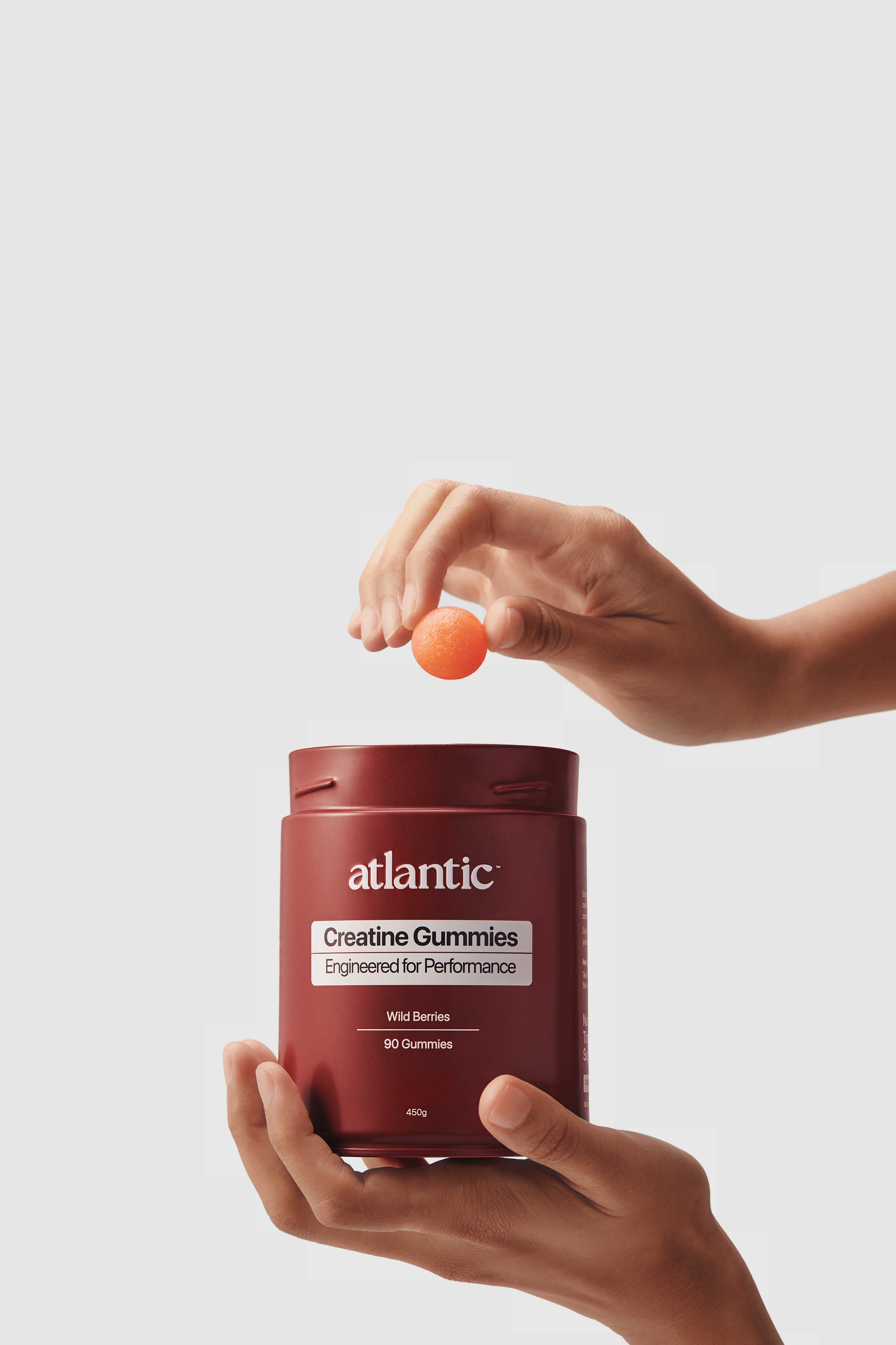 Hand holding a bottle of Atlantic Creatine Gummies with the label visible, a small orange gummy near the bottle, and a hand dropping a similar gummy into a container.
