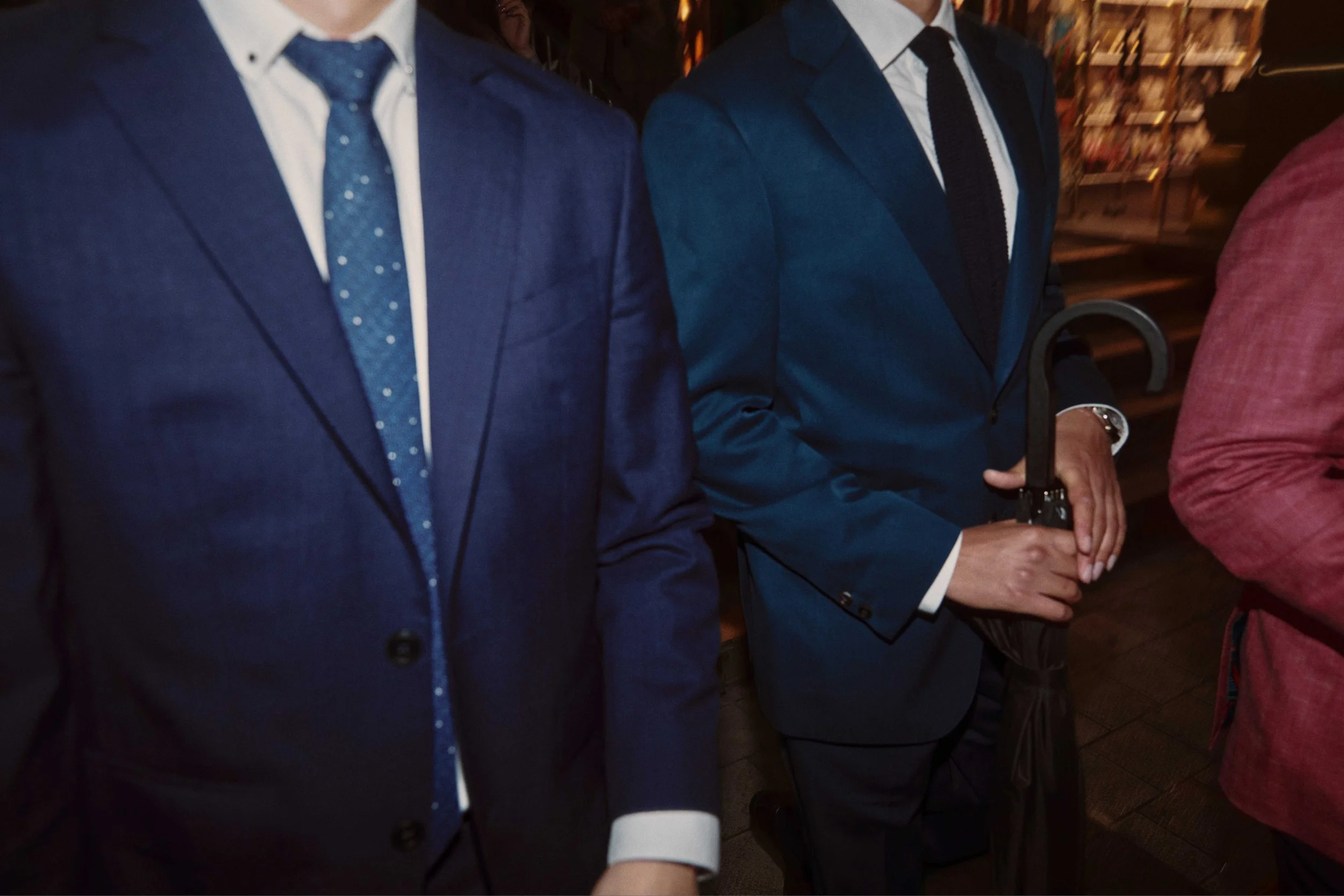 Close-up of people wearing formal suits, ties, and holding umbrellas in an indoor setting with shelves in the background.