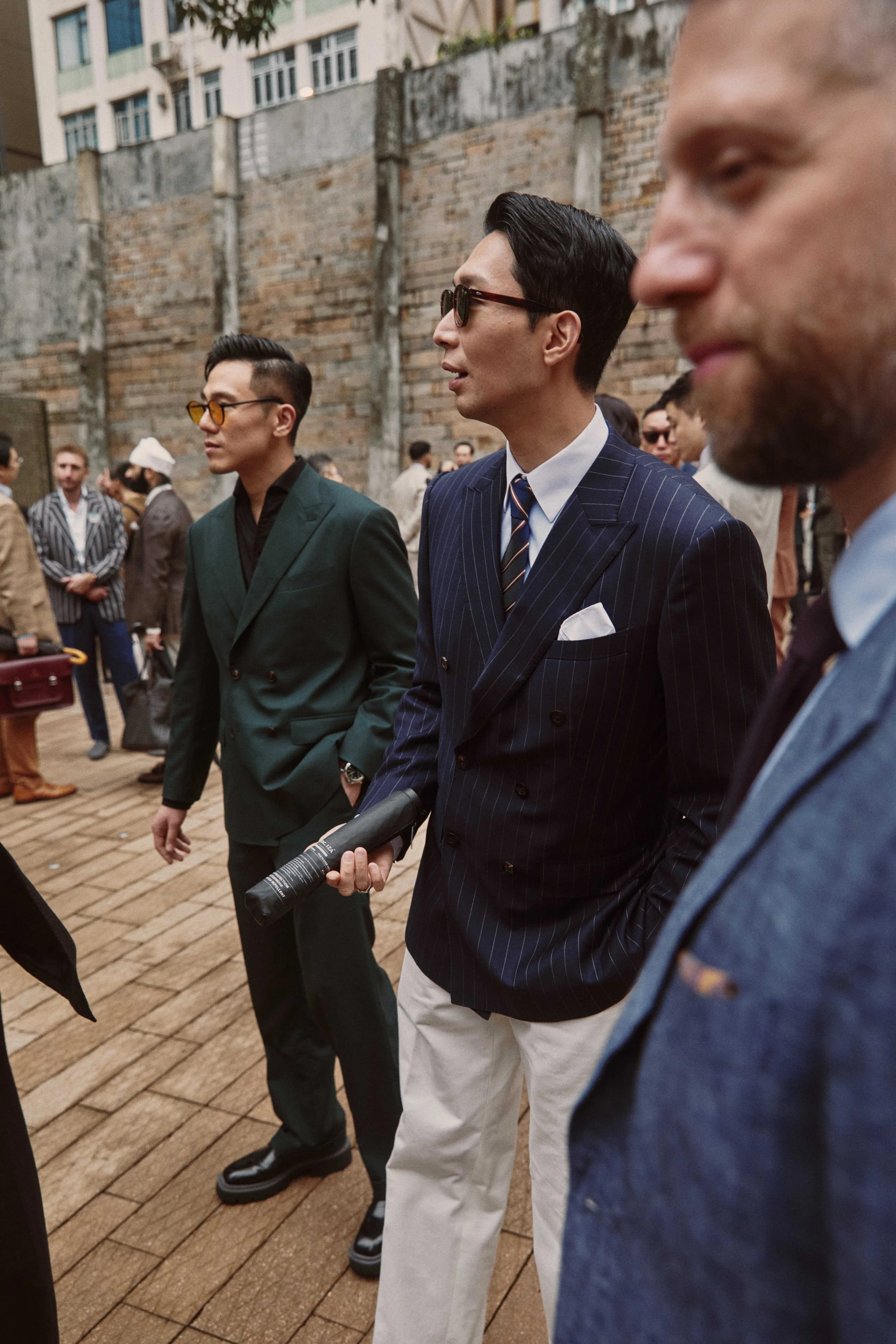 Group of men dressed in formal and casual attire walking outdoors near a brick wall and buildings, with some wearing sunglasses.