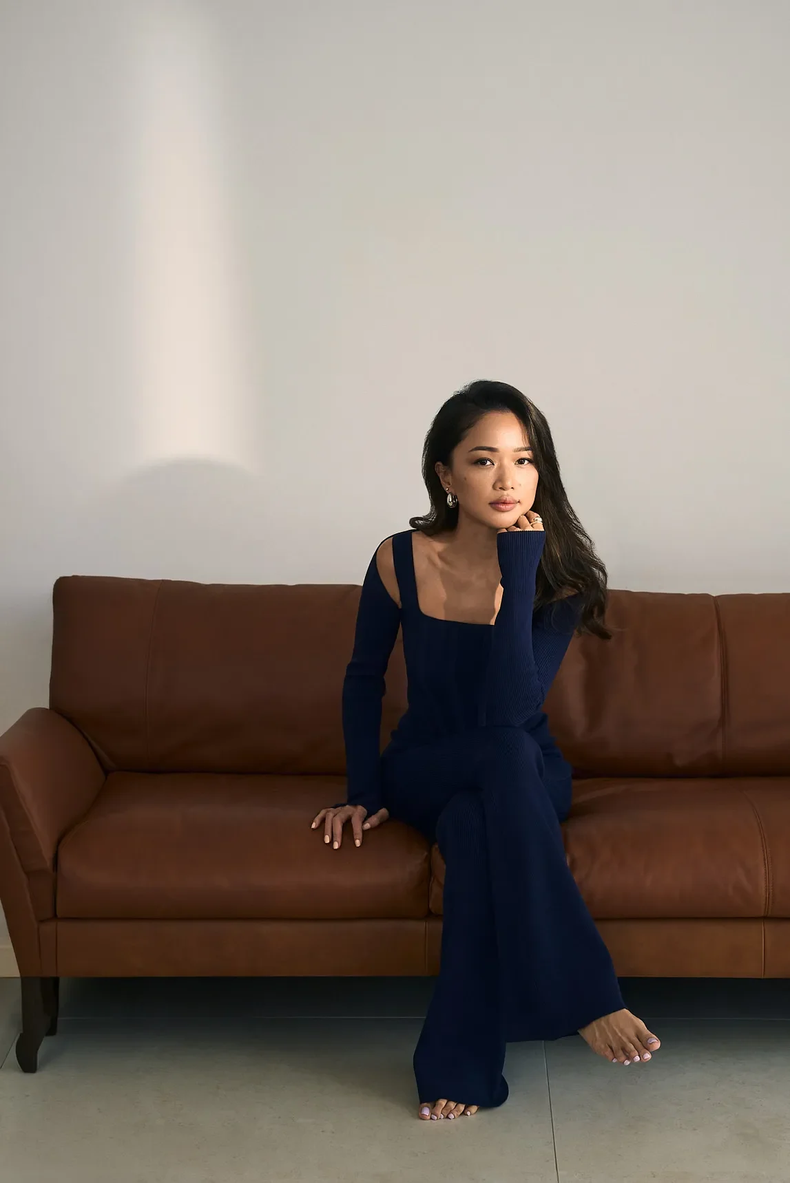 A woman with long dark hair and earrings is sitting on a brown leather sofa in a minimalist room with a plain white wall. She is wearing a navy blue outfit and is resting her chin on her right hand.