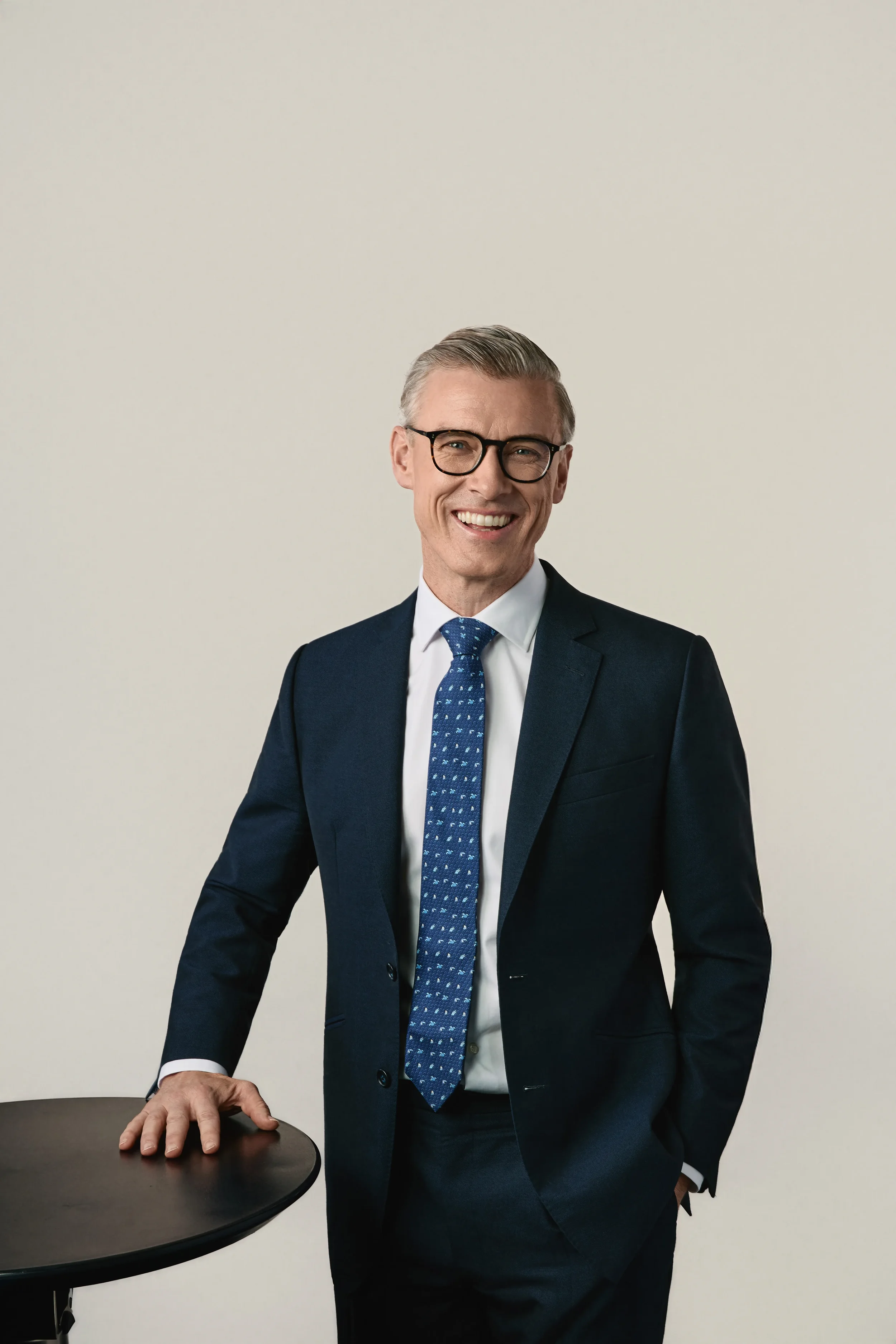 A smiling middle-aged man with gray hair and glasses wearing a navy blue suit, white shirt, and patterned blue tie, standing against a plain beige background with a small round table in front of him.