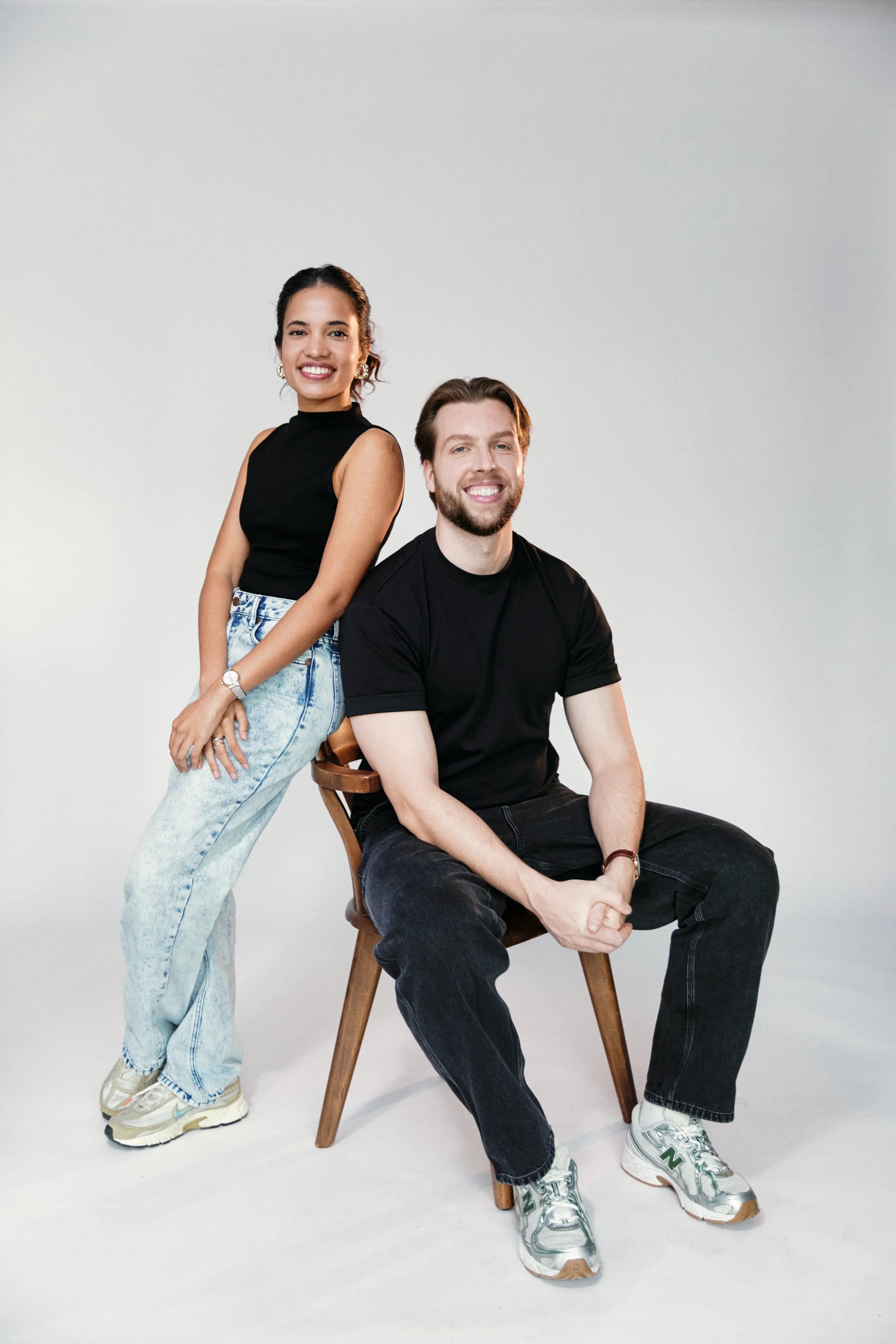 A young woman with dark hair in a ponytail wearing a black sleeveless top and light blue jeans standing next to a seated young man with brown hair, a beard, and a black t-shirt, both smiling, in front of a plain white background.