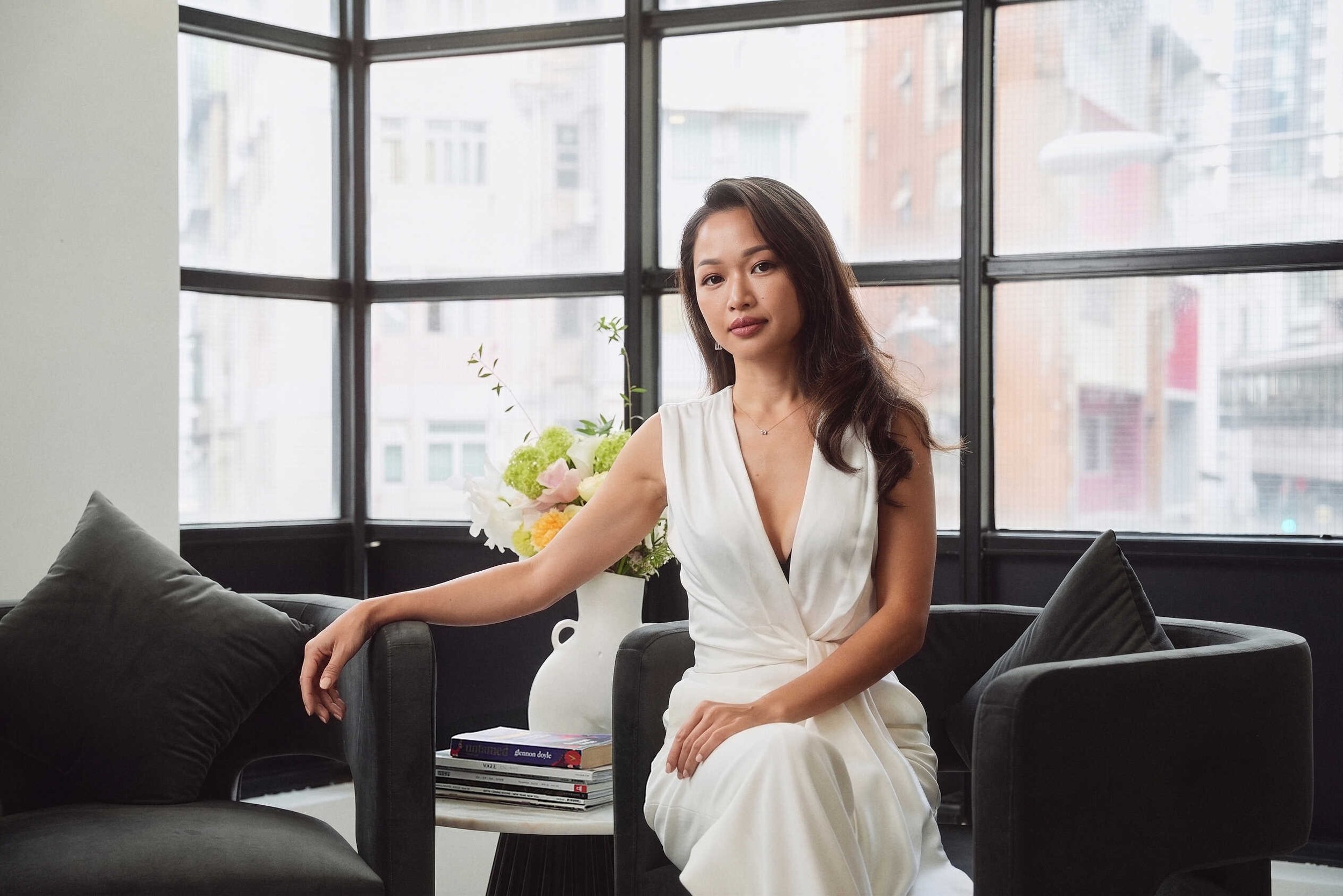 A woman in a white dress sitting on a black sofa in a modern living room with large windows, a bouquet of flowers behind her, and a stack of magazines on a small table.