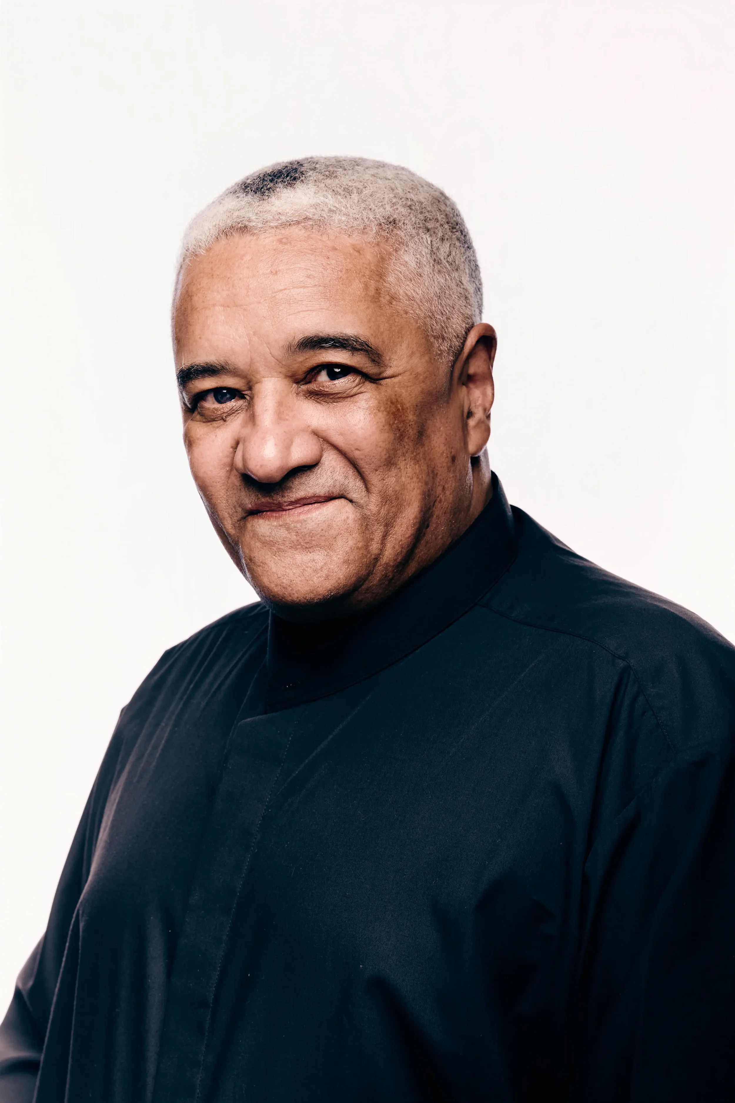 Close-up of a middle-aged man with short, gray hair, wearing a black, high-collared shirt, against a plain white background.