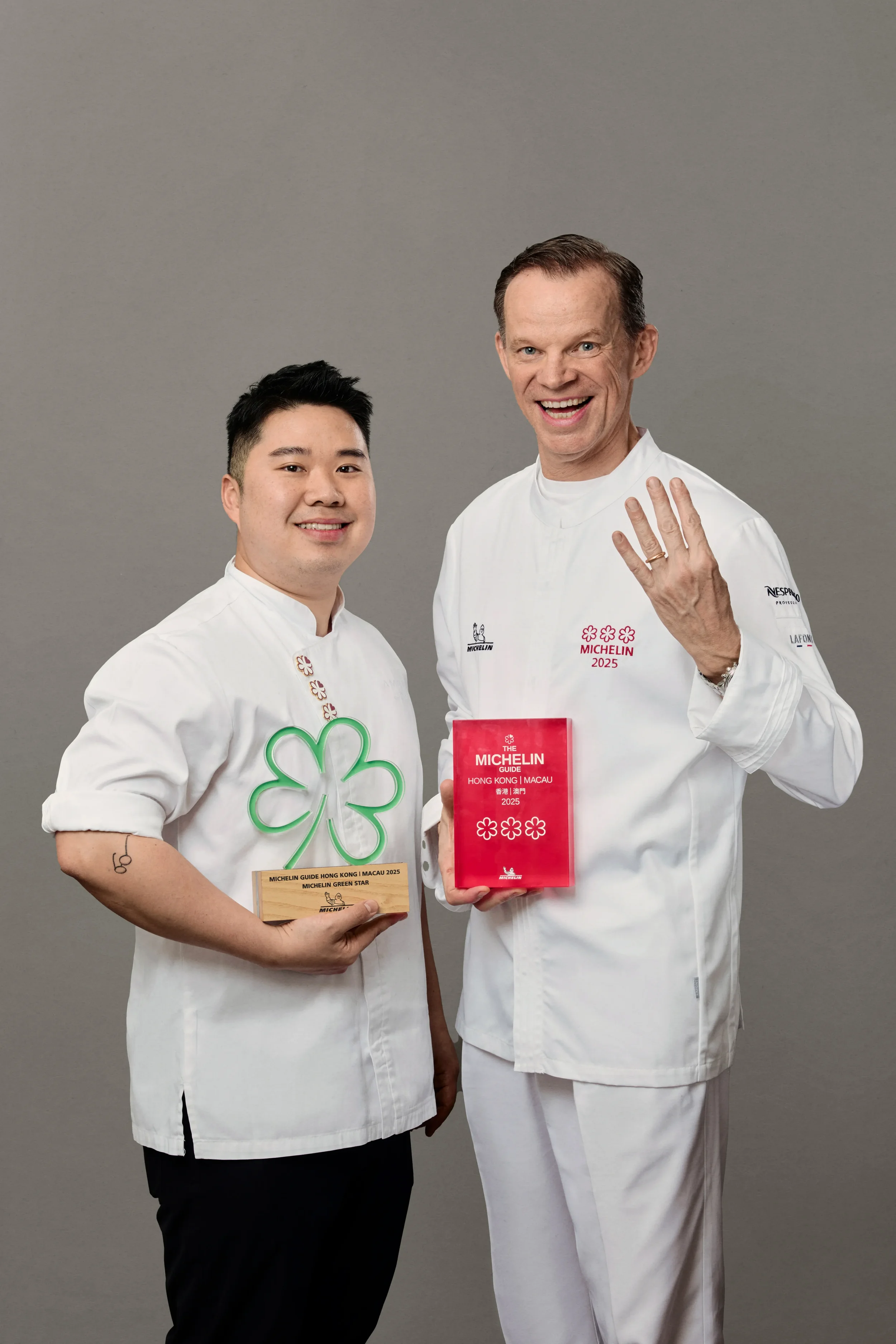 Two male chefs stand side by side, smiling, against a plain gray background. The chef on the left holds a wooden plaque reading 'Michelin Guide Hong Kong | Macau 2025 Michelin Green Star.' The chef on the right raises a hand while holding a red box r
