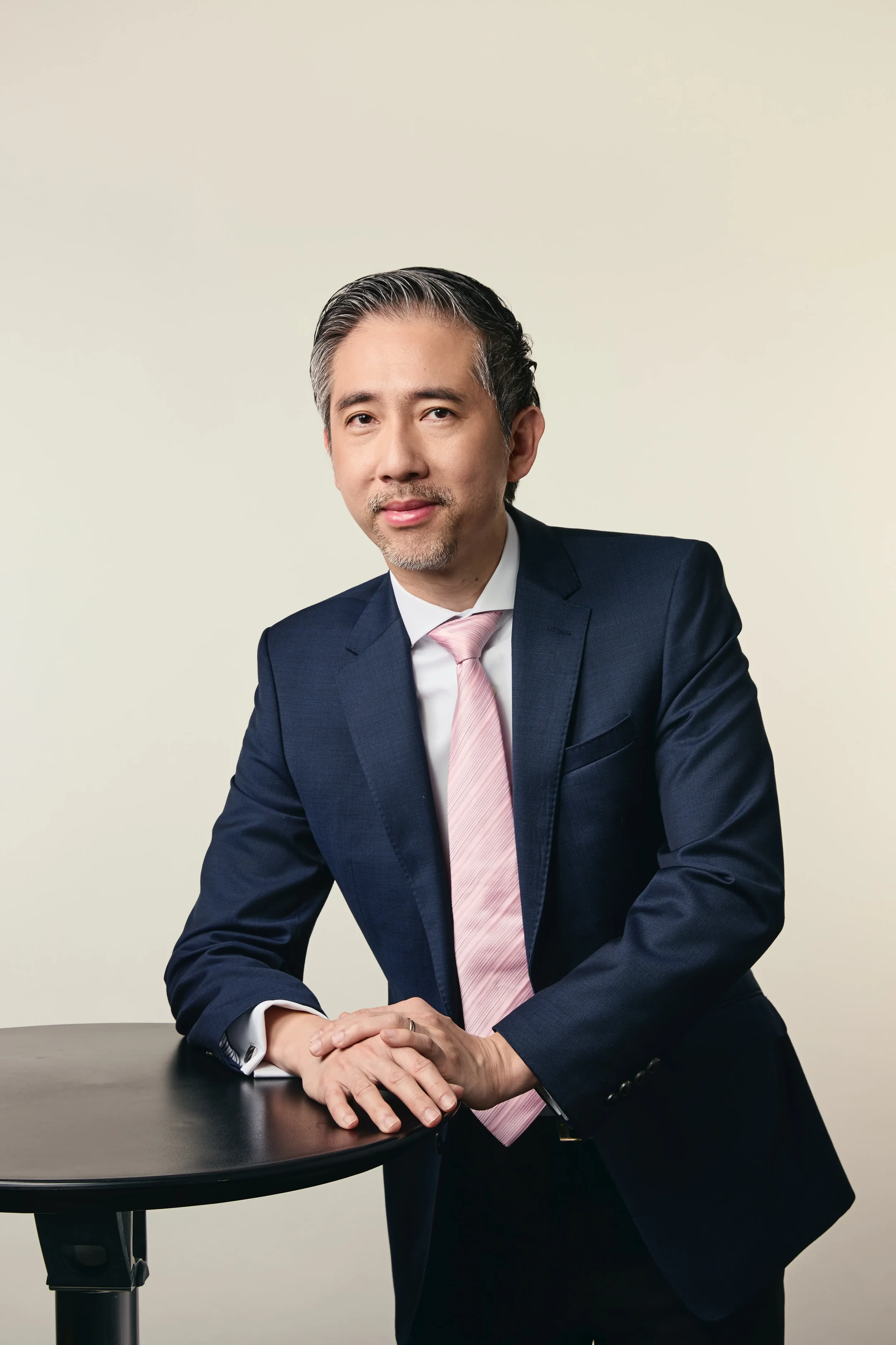 A man in a dark suit with a pink tie, sitting at a black table with hands clasped, against a plain light-colored background.
