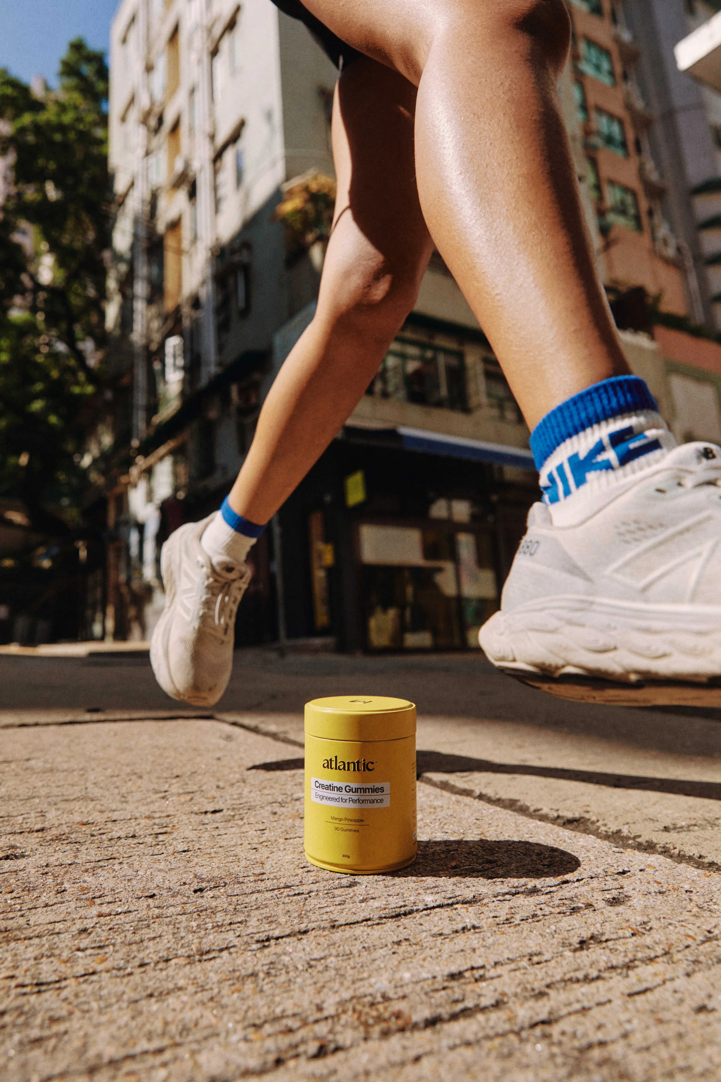 Close-up of a person jumping in athletic shoes with blue and white socks, on a city street with apartment buildings in the background. A yellow container labeled 'Atlantic Creatine Gummies' is on the ground.