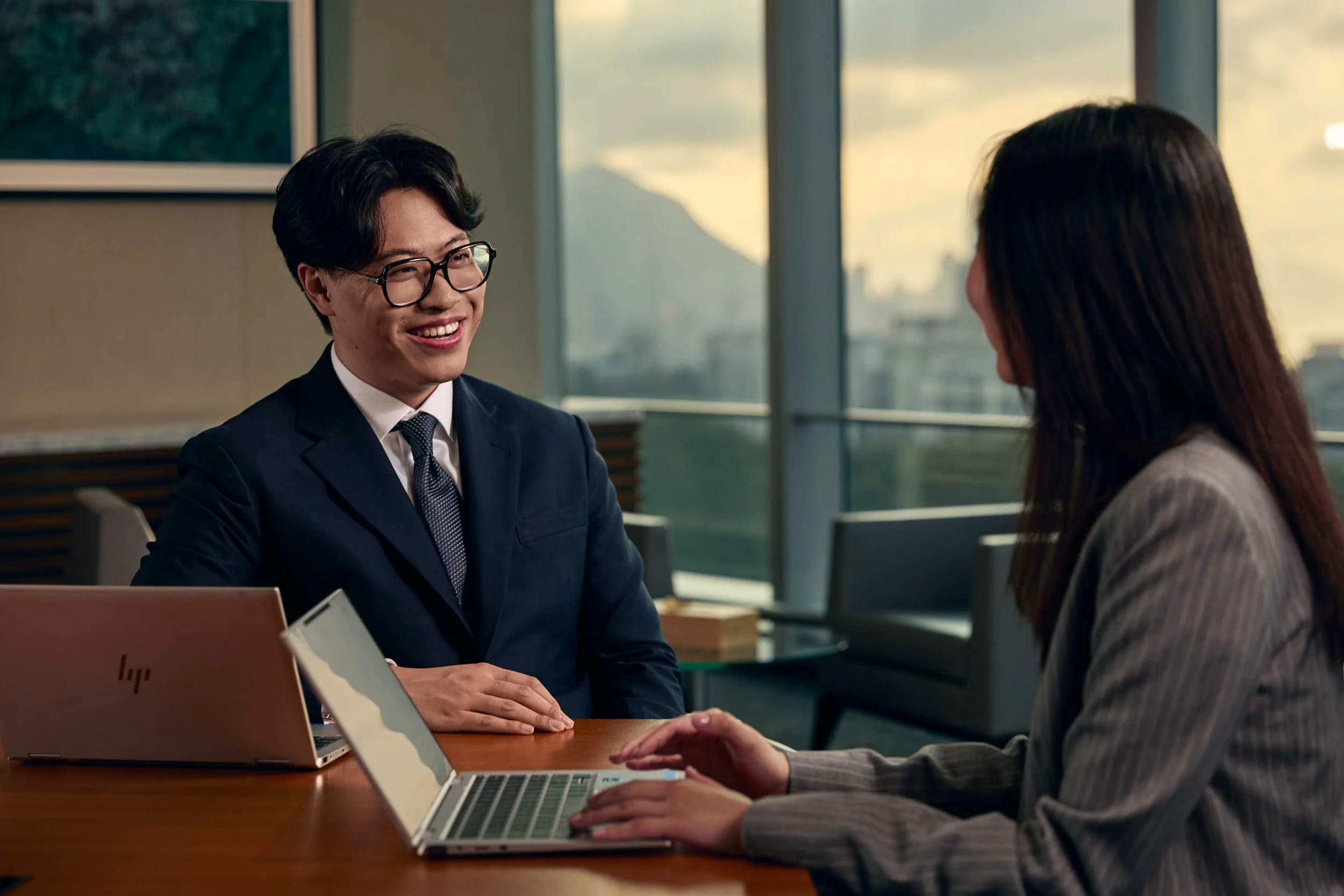 A man in a suit and glasses smiling while talking to a woman in a business meeting, both seated at a wooden table with laptops, in a modern office with large windows showing a cityscape at sunset.