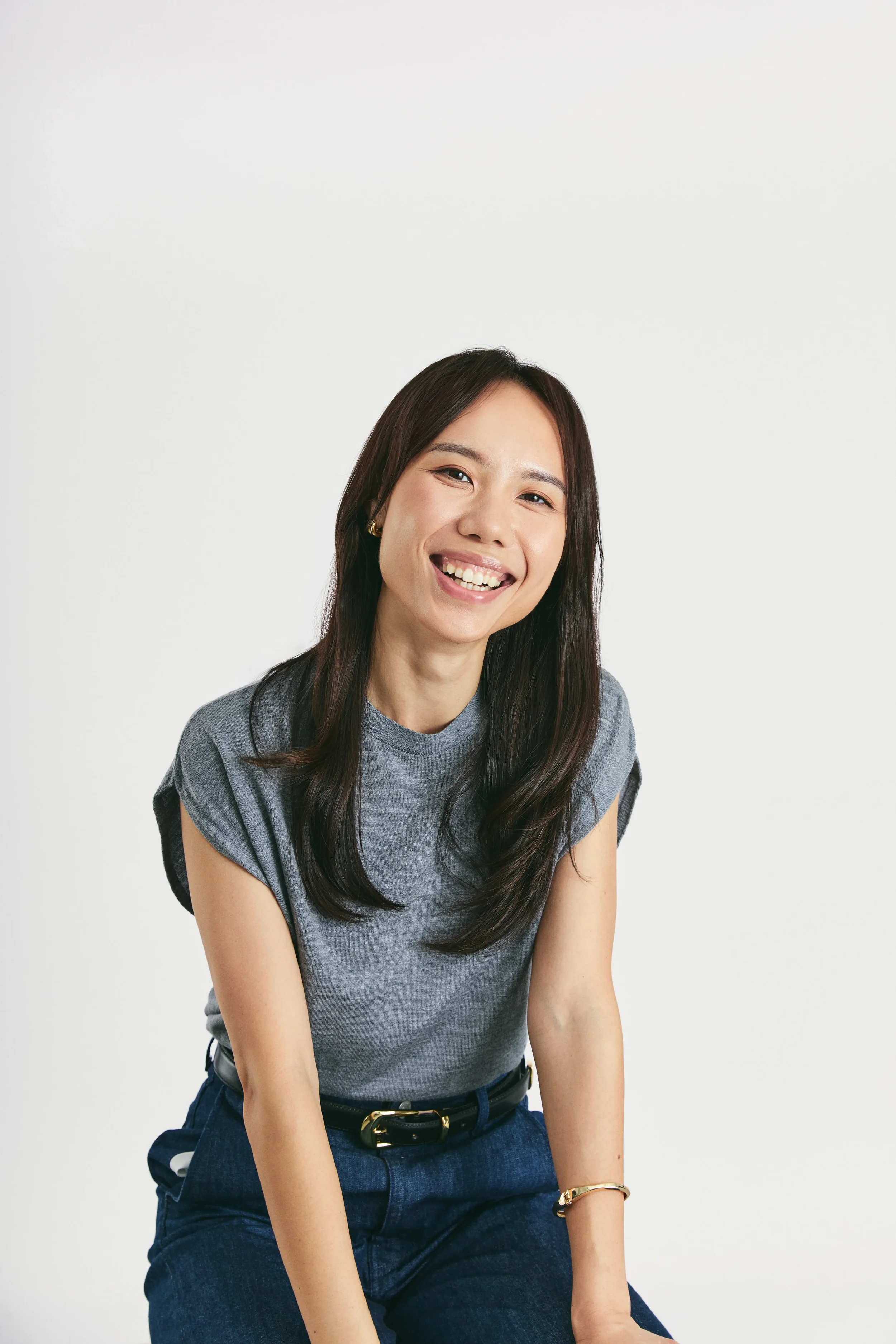 A woman with long dark hair smiling at the camera, wearing a gray t-shirt and dark blue jeans with a black belt, in front of a plain white background.
