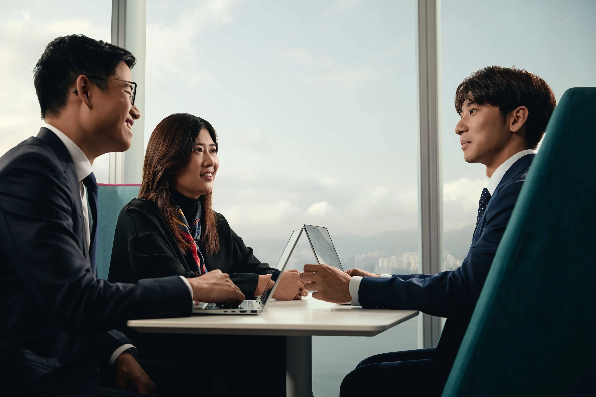 Four business professionals, two men and two women, sitting at a conference table engaged in a discussion. They are dressed in business attire and appear to be in a high-rise office with a large window revealing a cityscape and partly cloudy sky.