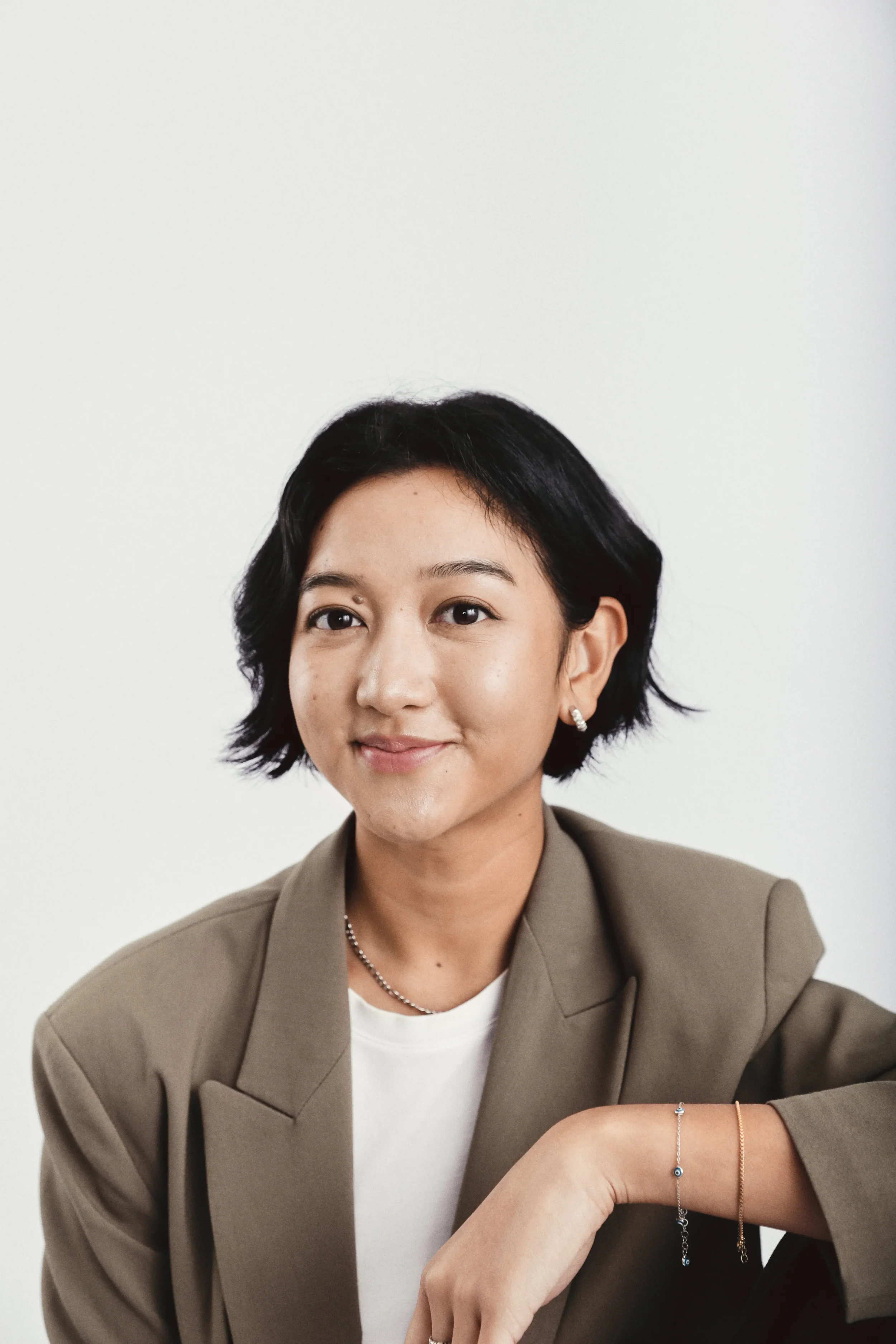 A woman with short black hair wearing a beige blazer, white t-shirt, and jewelry, sitting at a table with a neutral white background.