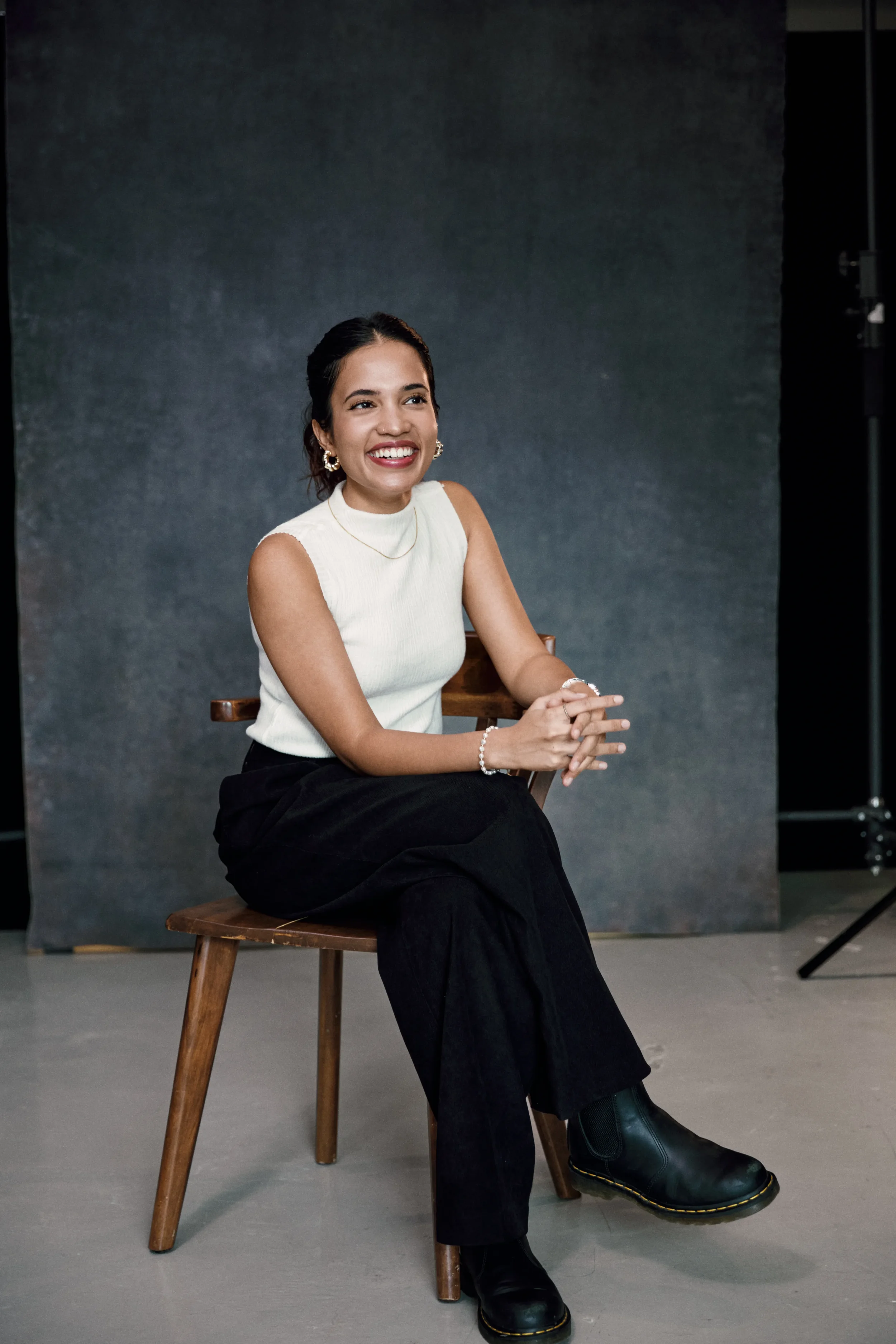 A woman sitting on a wooden chair, smiling and looking to her right, wearing a sleeveless white top, black pants, and black boots, in a studio setting with a dark backdrop.