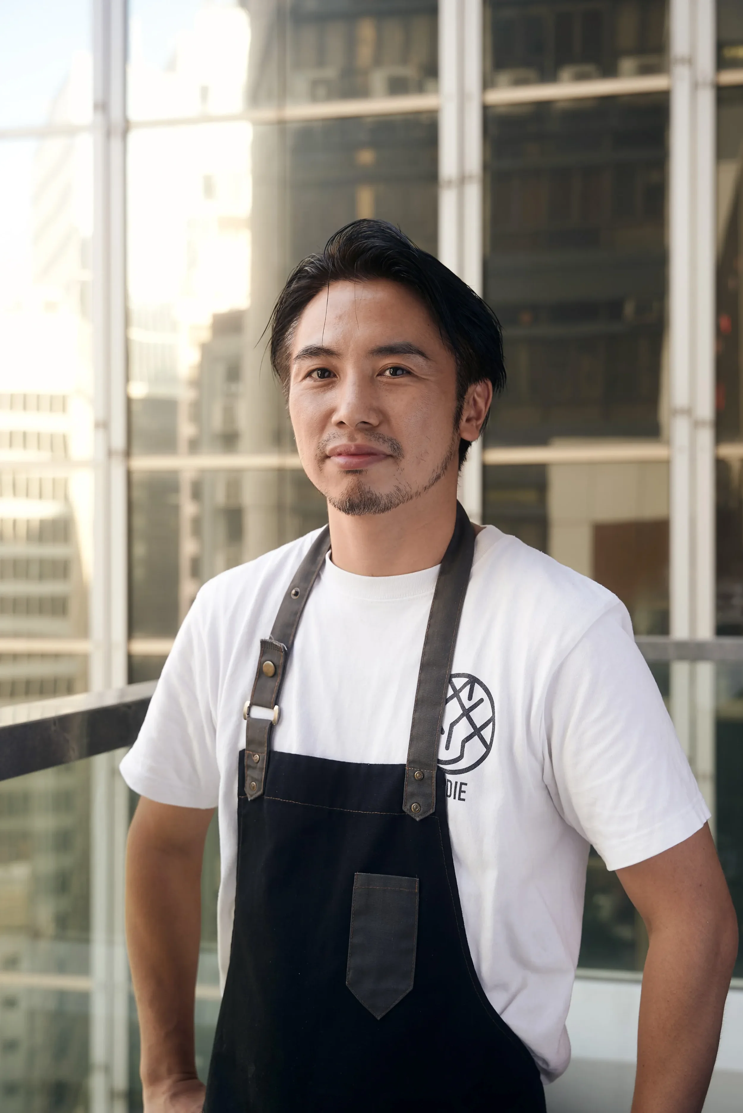 A young man with dark hair and facial hair stands outside a glassed building, wearing a white T-shirt and a black apron with a logo, looking at the camera.