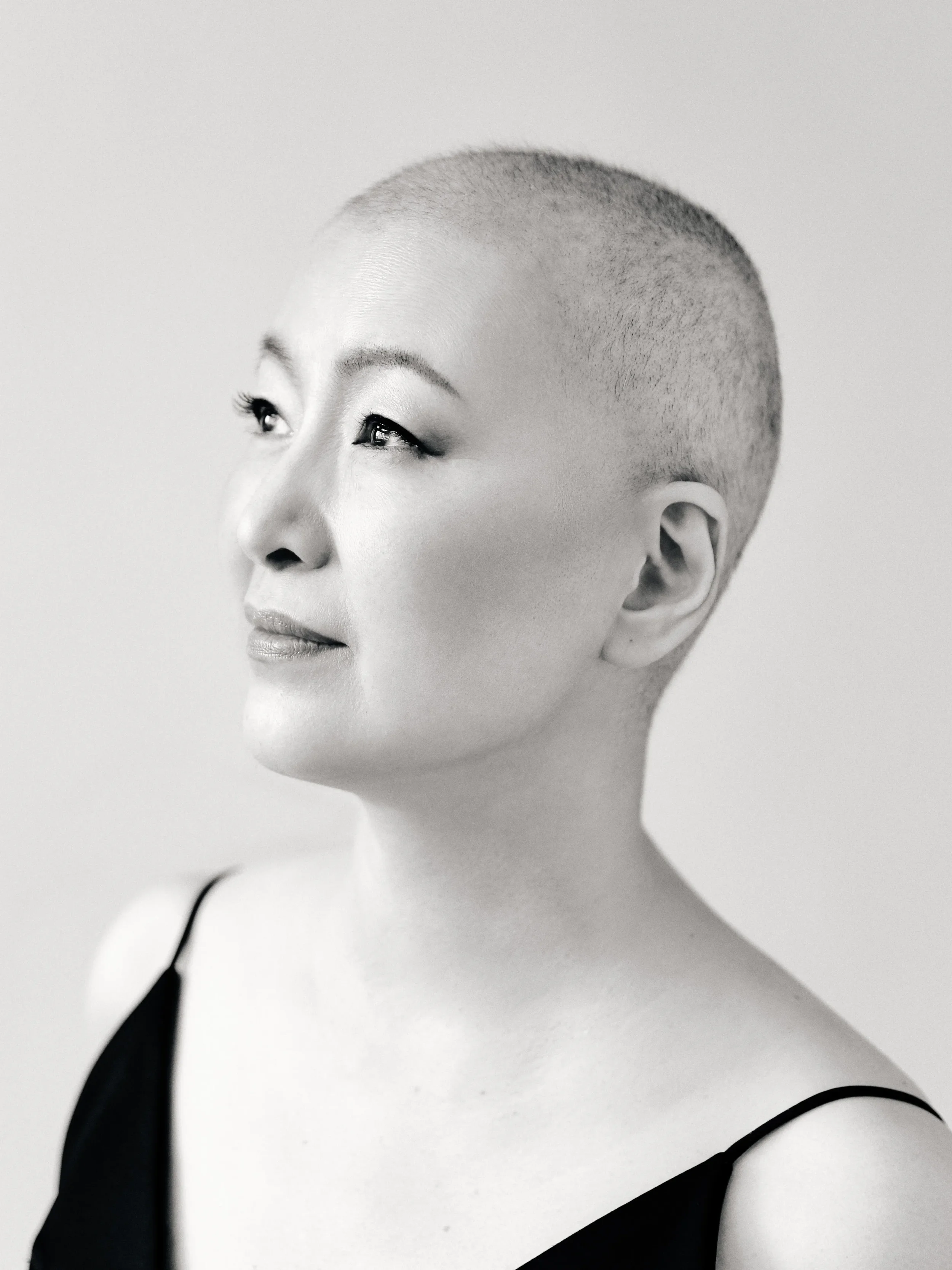 A black and white photograph of a woman with a short buzz haircut, looking slightly upward and to her right, wearing a thin-strapped top.