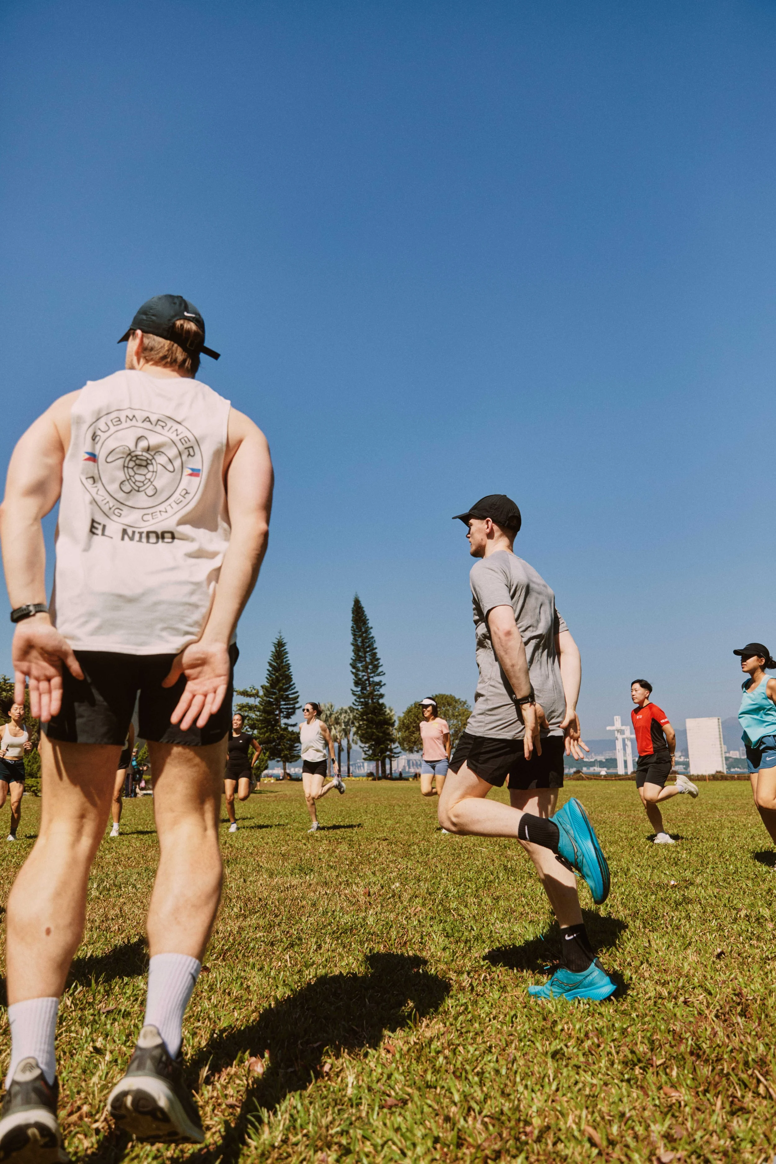 People participating in an outdoor running event or exercise class on a grassy field, with some individuals kneeling and others standing under a clear blue sky.