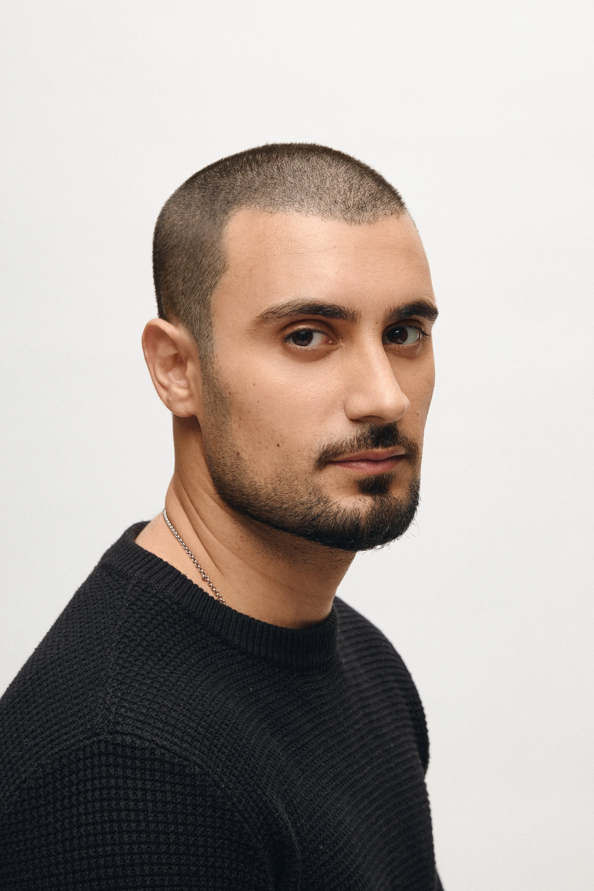 Close-up of a Zaref Khan with a buzz cut hairstyle, wearing a black sweater and a silver chain necklace, against a plain white background.