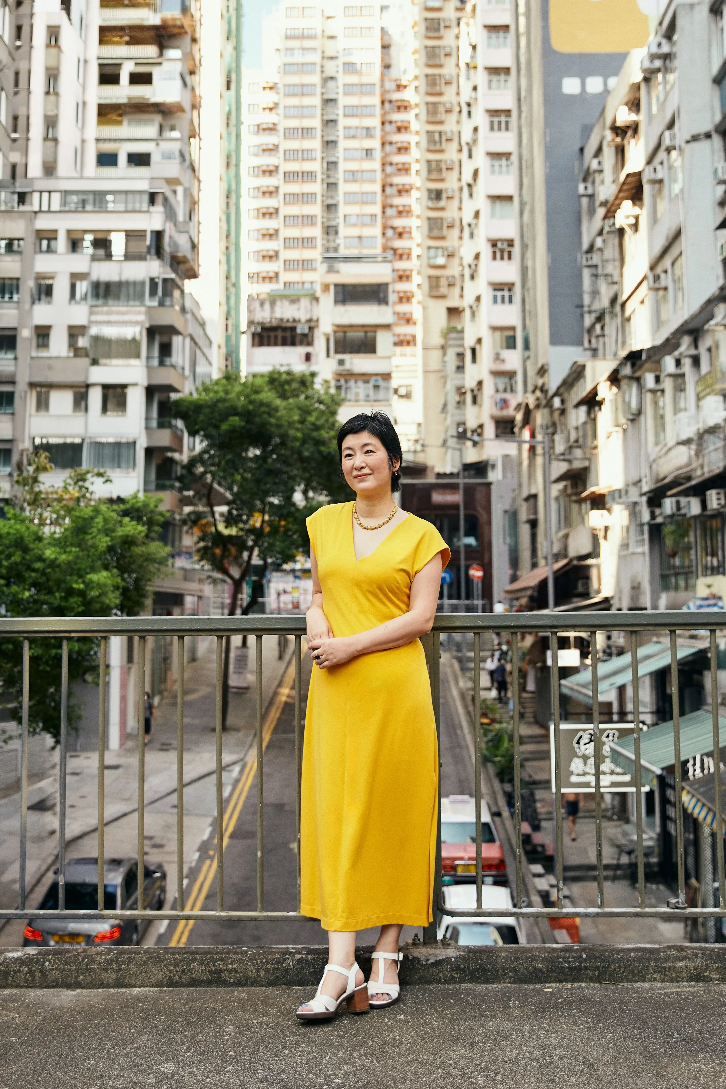 A woman in a yellow dress and white high-heeled sandals standing outdoors on a balcony or elevated platform, with a city street and tall apartment buildings in the background.