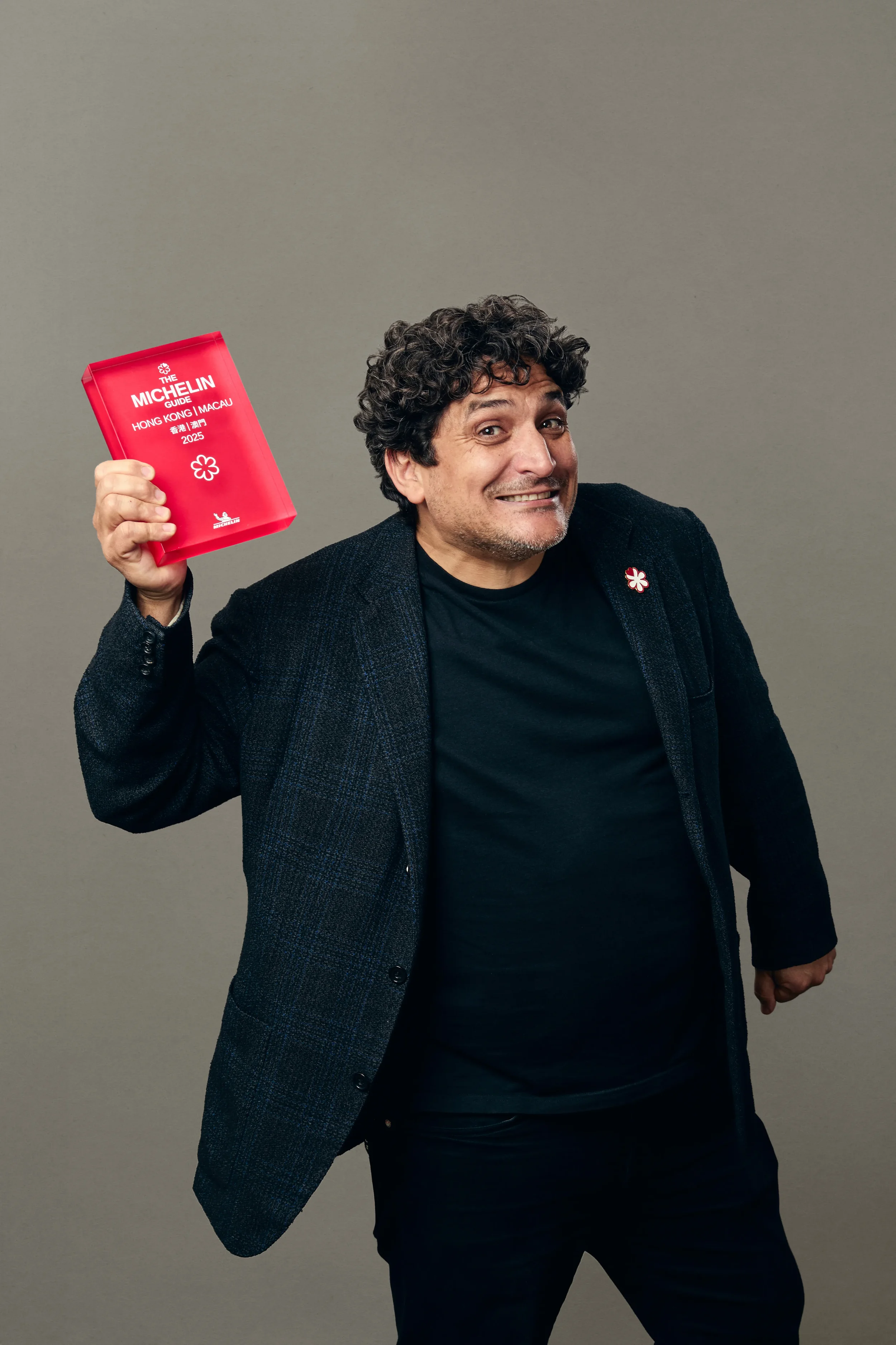 Man smiling, dressed in black shirt and dark blazer, holding a red Michelin guidebook for Hong Kong and Macau 2025, standing against a plain gray background.