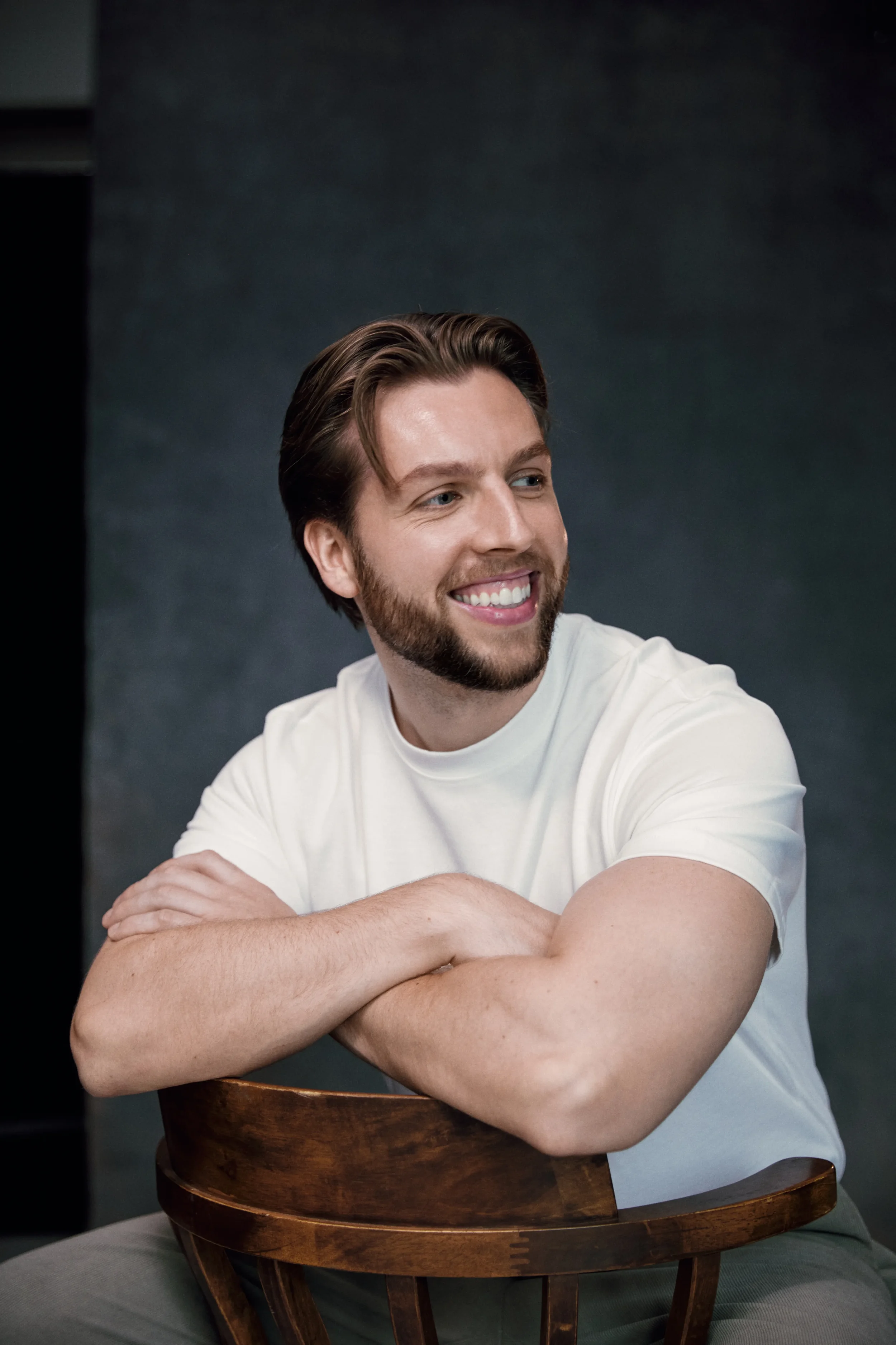 A smiling man with brown hair and a beard, wearing a white t-shirt, leaning on a wooden chair with his arms crossed, in front of a dark background.