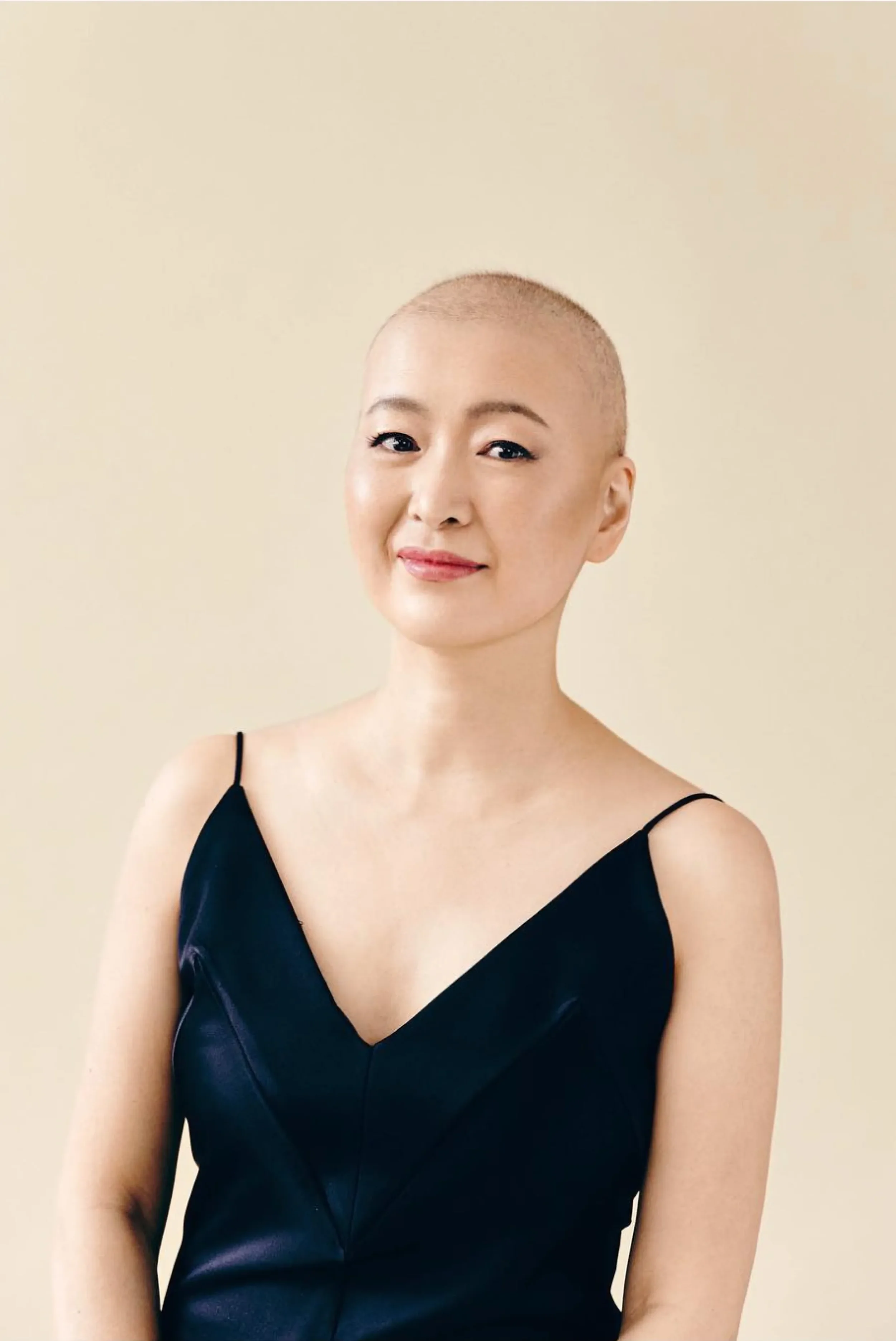 A woman with a shaved head, smiling, wearing a black spaghetti strap dress, against a plain beige background.