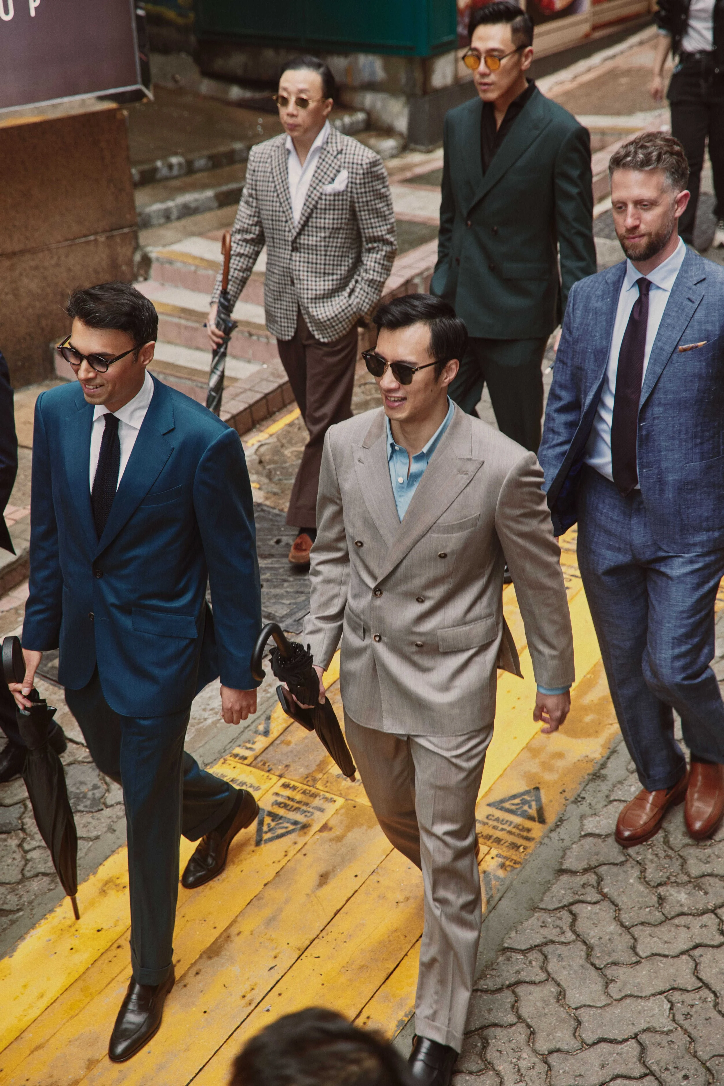 A group of well-dressed men and women walking together on a city sidewalk, some wearing sunglasses and suits.
