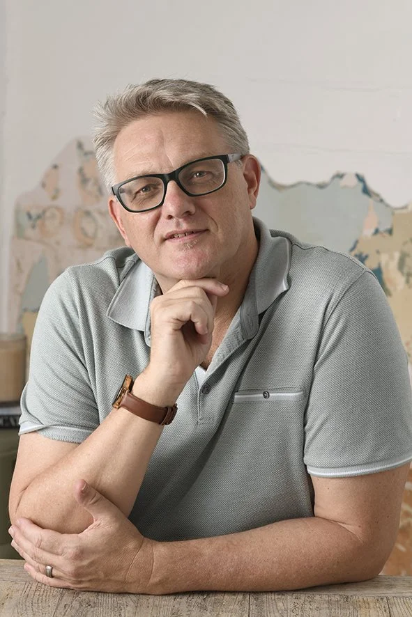 A middle-aged man with gray hair and glasses, wearing a gray polo shirt, sitting at a table with one arm crossed and the other hand resting on his chin, indoors with a painted wall in the background.