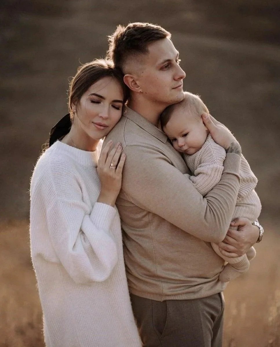 A family of three embracing outdoors during sunset, with a woman resting her head on a man holding a young child.