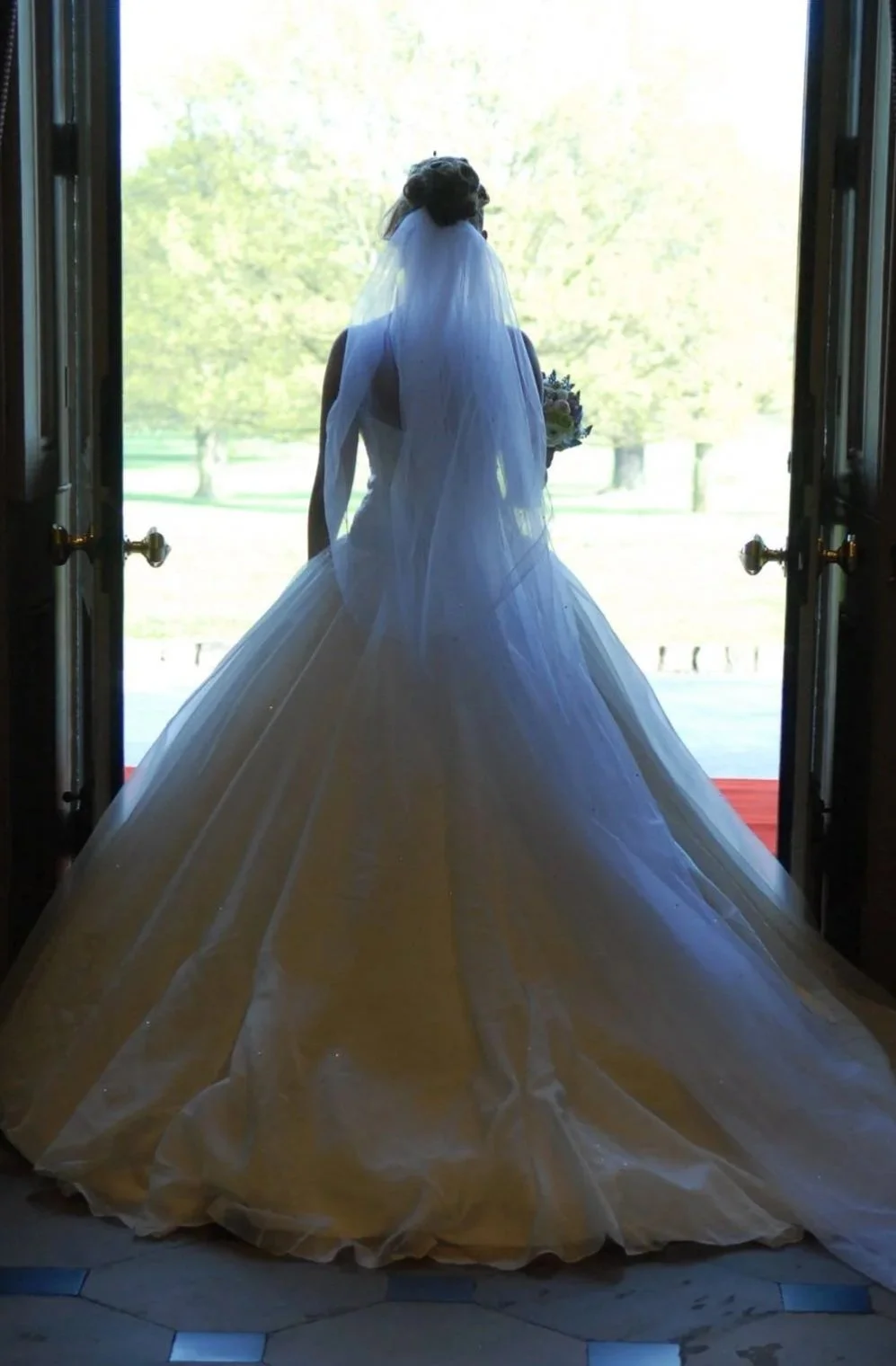 Bride in a wedding gown with a veil, standing in a doorway overlooking a green outdoor scene, holding a bouquet of flowers.