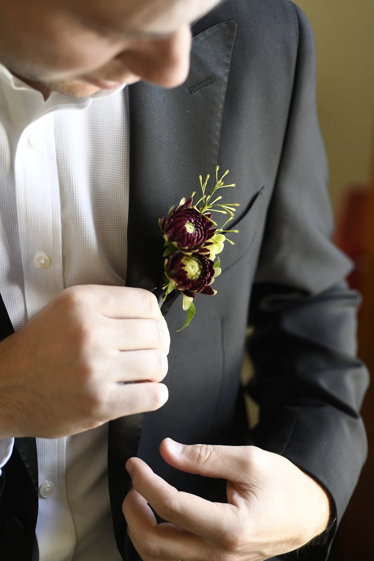A man in a black suit and white shirt pinning a boutonniere with dark purple and yellow flowers on his left lapel.