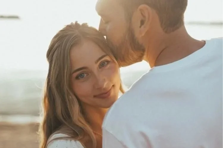 A woman with long blonde hair smiling as a man kisses her on the cheek at the beach during sunset.