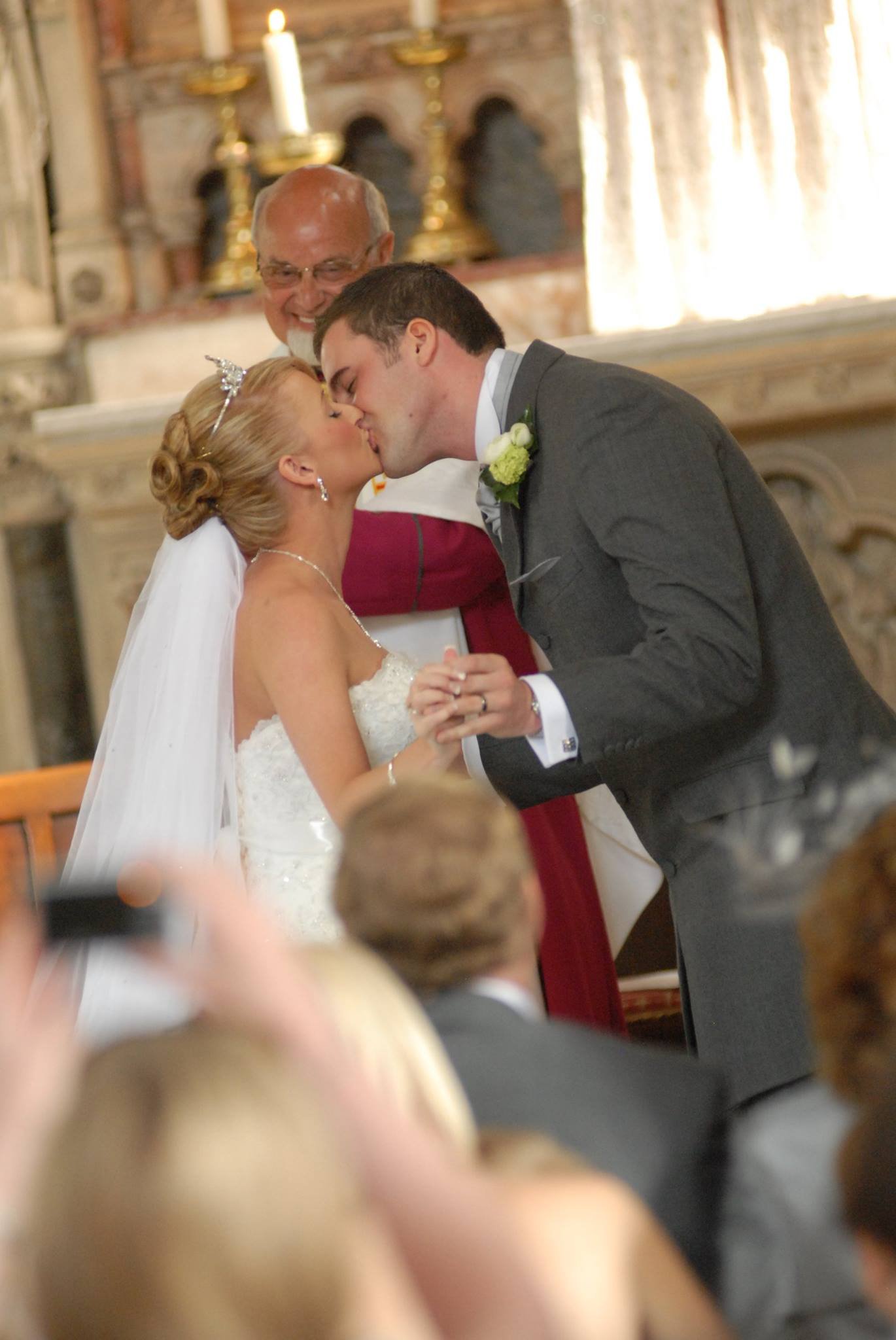 A bride and groom kiss during their wedding ceremony inside a church, with an officiant smiling behind them, in front of an altar.