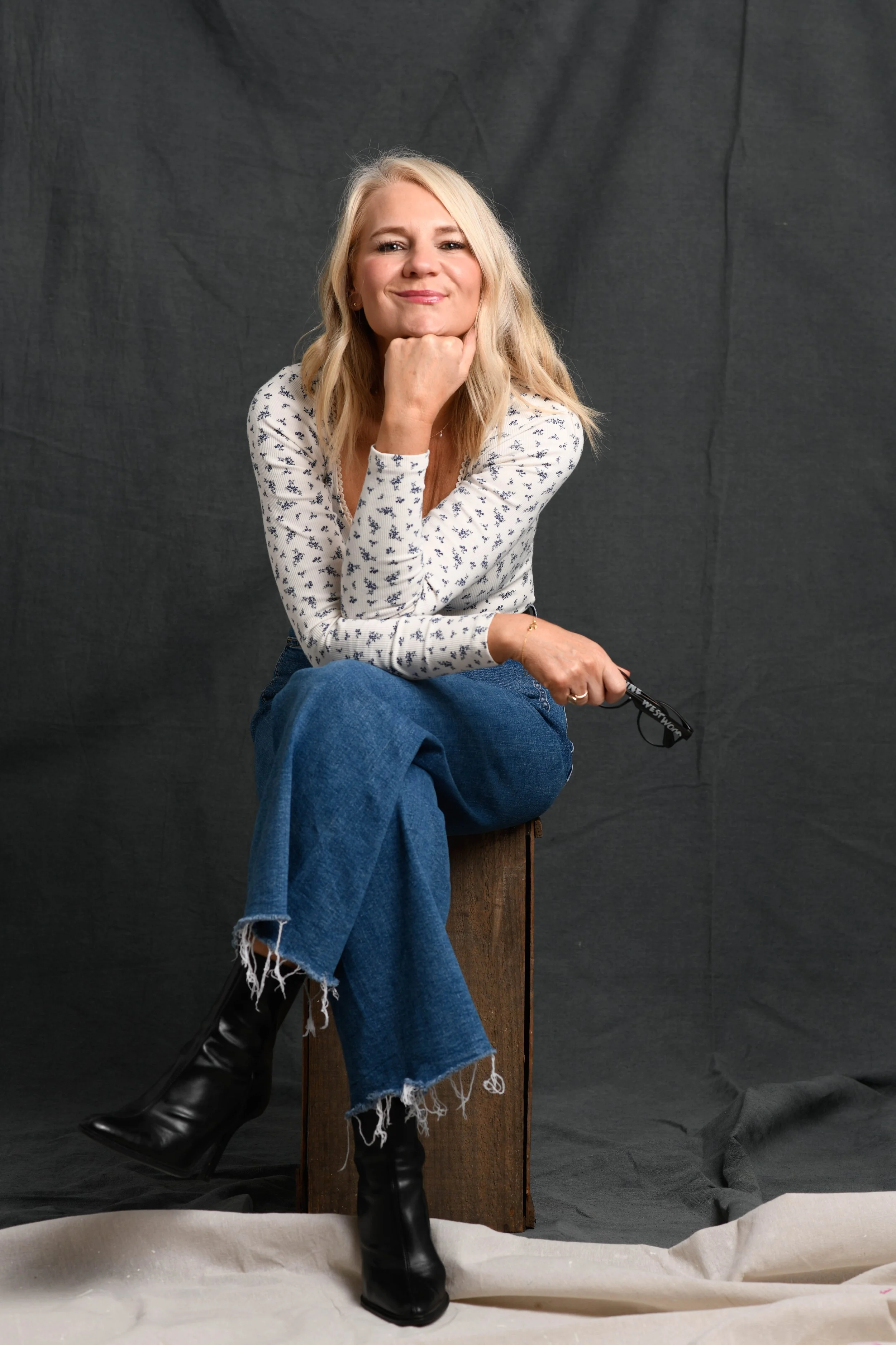 A woman with blonde hair, wearing a white shirt with a small black pattern, blue jeans with frayed hems, and black boots, sitting on a wooden stool against a plain dark background. She is resting her chin on her hand and smiling at the camera.
